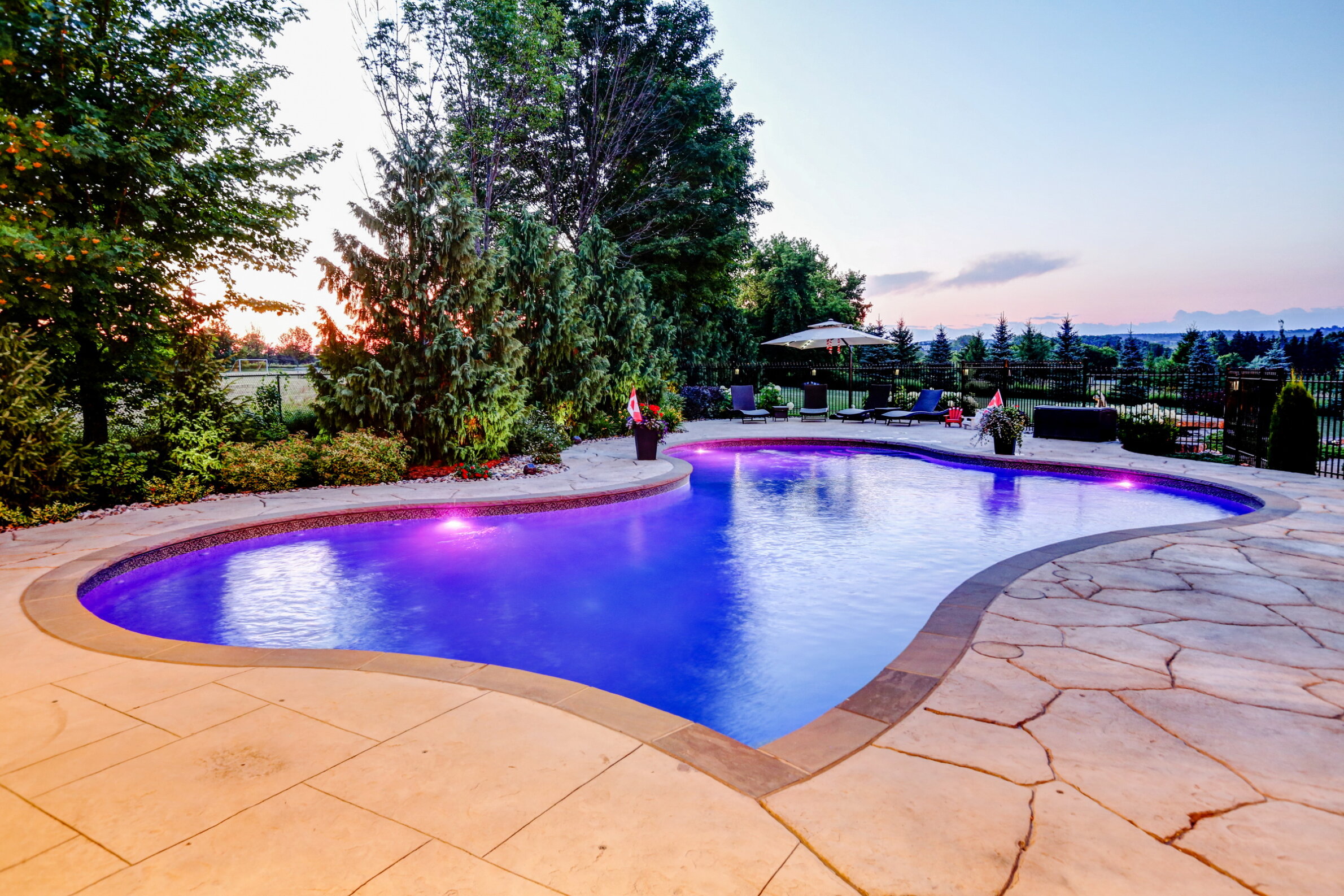 A luxurious outdoor swimming pool with purple lighting at dusk, surrounded by trees, a patio with lounge chairs, and a gazebo.