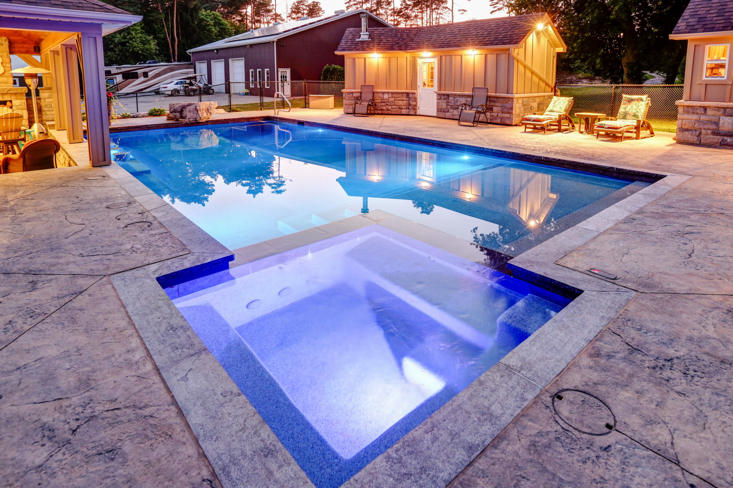 A luxurious residential swimming pool at dusk with clear blue waters, adjacent spa, patio furniture, and a house with warm lighting in the background.
