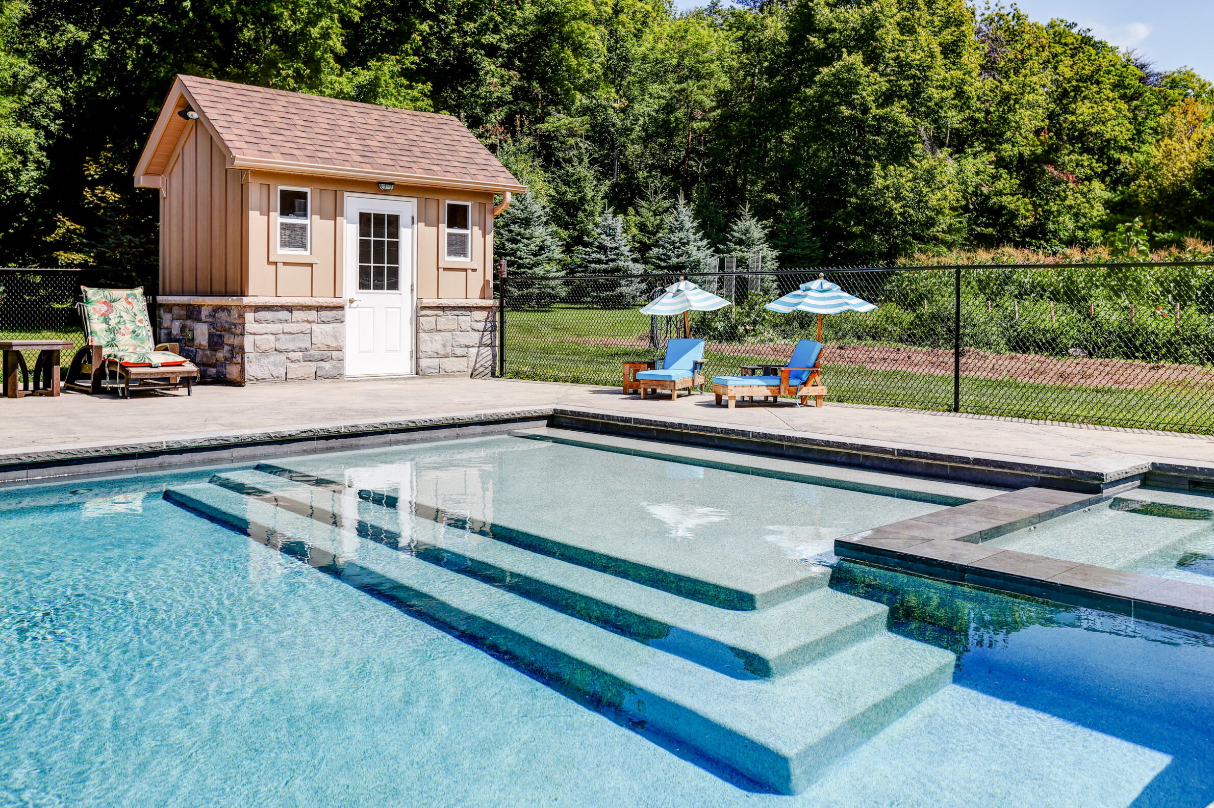 Outdoor swimming pool with clear blue water, steps leading in, surrounded by a patio, lounge chairs, umbrellas, and a small pool house, trees in the background.