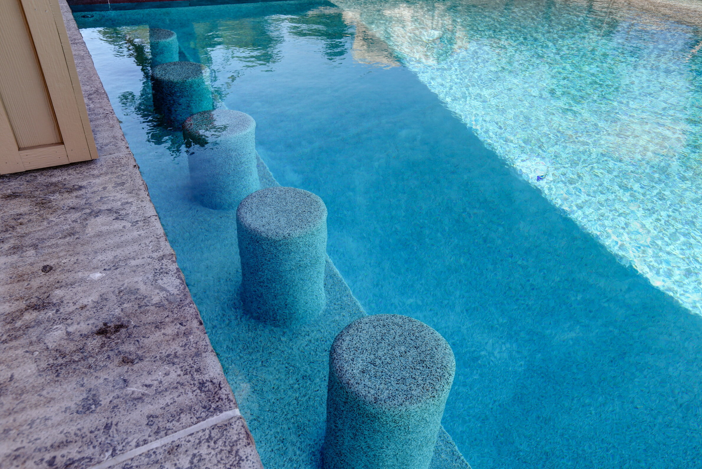 A series of rounded stone steps lead across the clear, blue water of a tranquil swimming pool adjacent to a concrete patio.