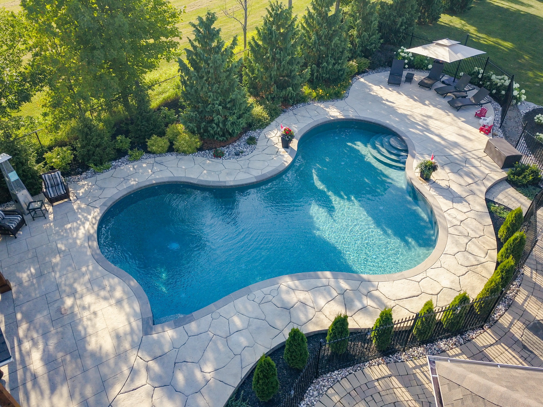 An aerial view of a residential backyard featuring a kidney-shaped swimming pool, patio with furniture, umbrellas, neatly landscaped garden, and trees.