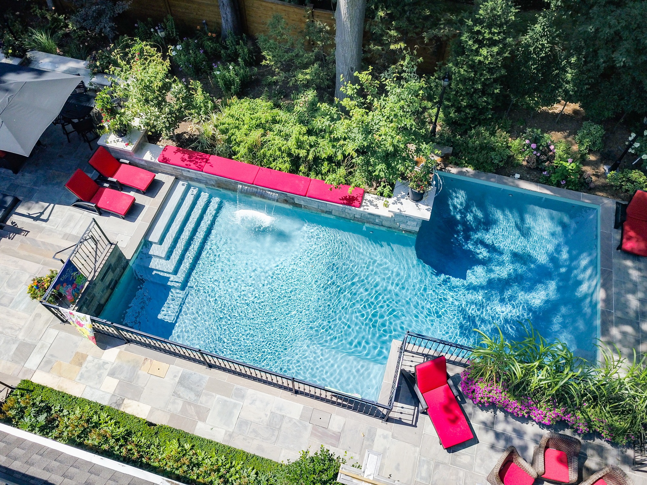 An aerial view of a backyard with a rectangular pool, surrounded by loungers, landscaping, and a patio area, on a sunny day.