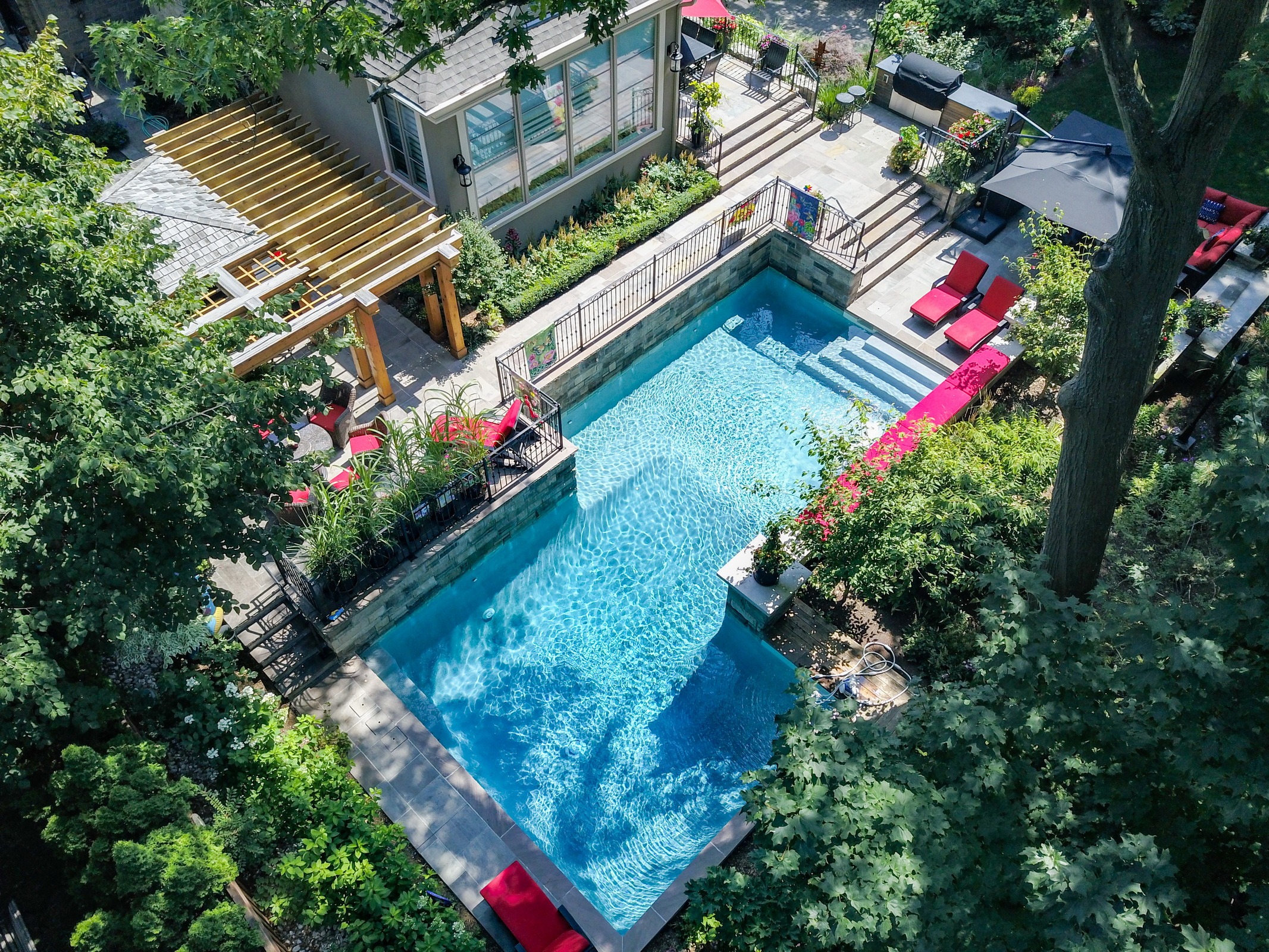 Aerial view of a backyard with a rectangular pool, surrounded by lush greenery, patio furniture, and a pergola next to a house.
