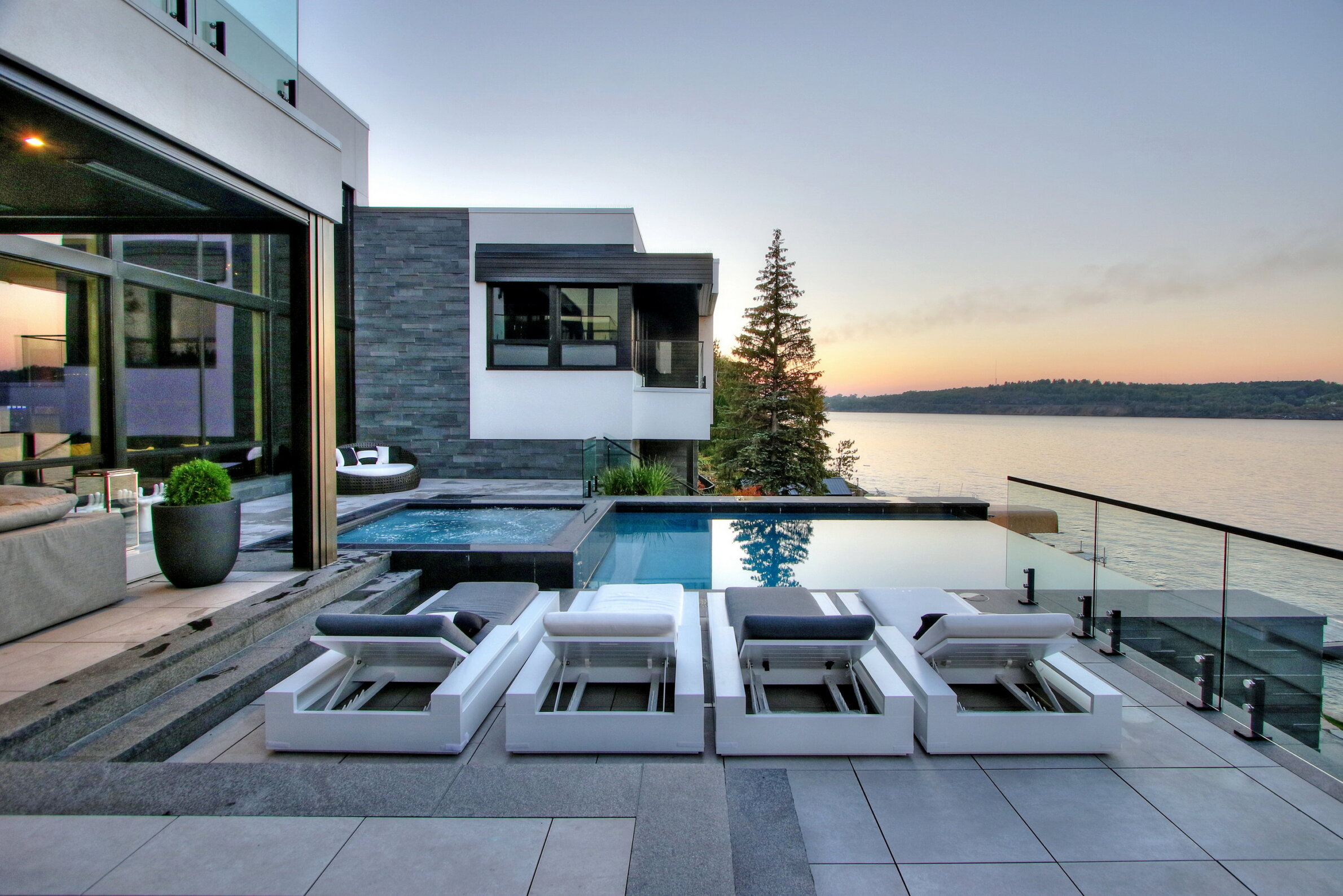 Modern house with infinity pool overlooking a tranquil body of water at dusk. The terrace includes lounge chairs and a seamless indoor-outdoor living space.