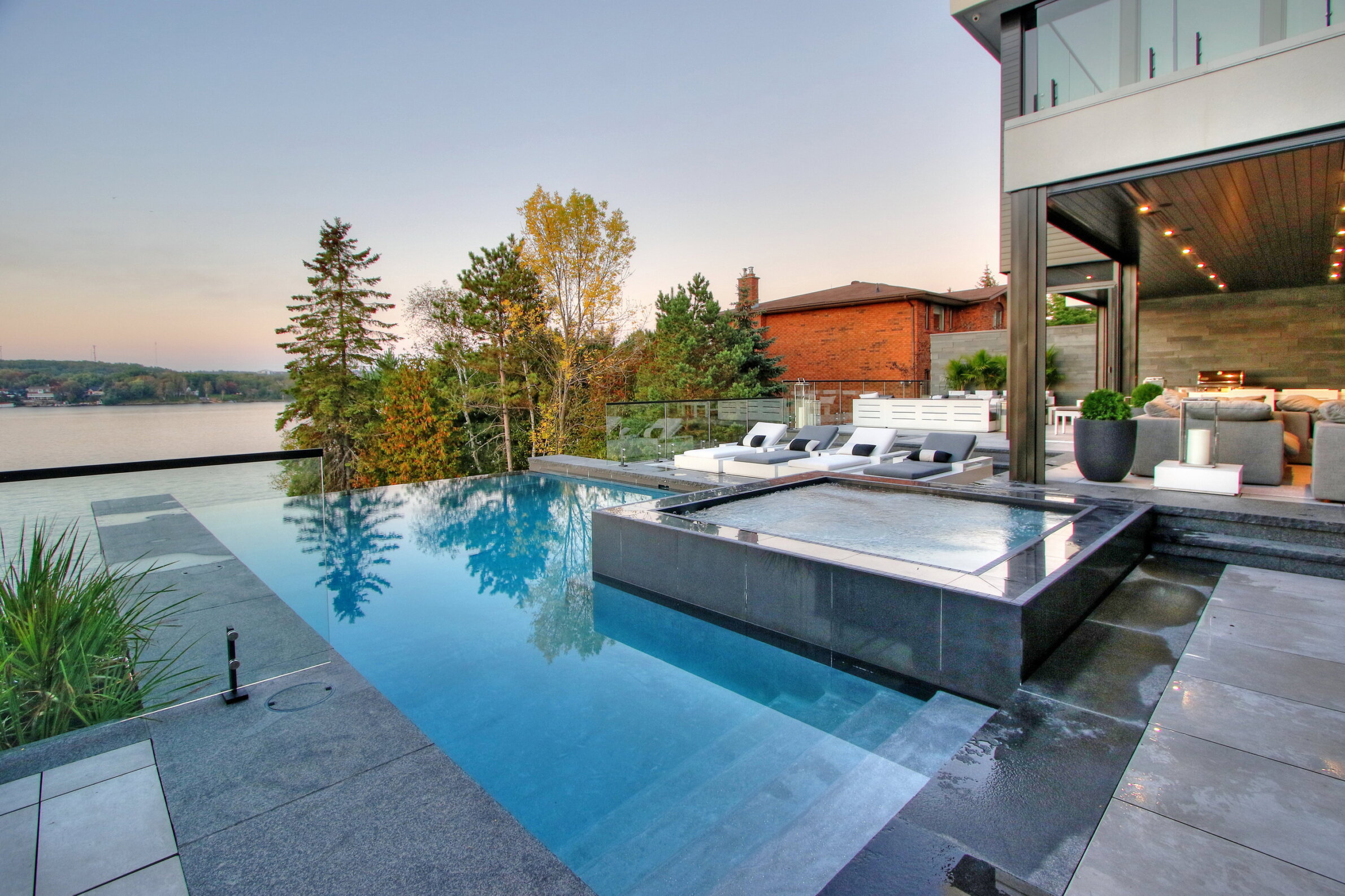 An outdoor infinity pool blending with a lake view, adjacent hot tub, loungers, and a cozy seating area with a fireplace. Modern luxury home setting.