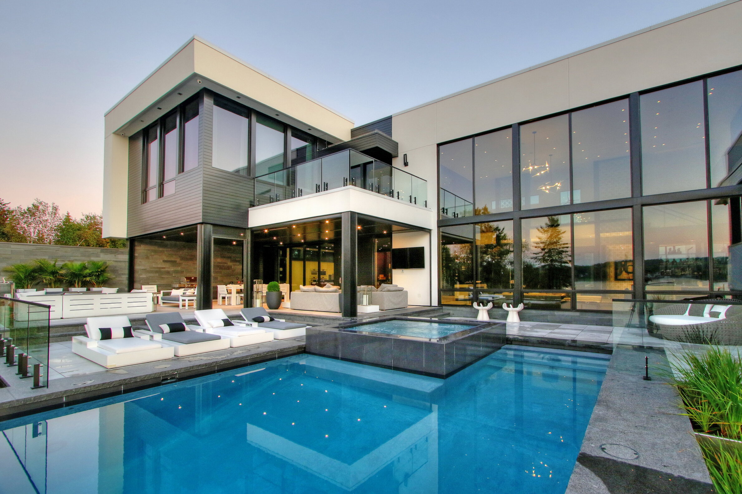 A modern two-story house with large windows, an outdoor pool, patio furniture, and a clear sky at dusk. Luxurious and sleek architecture.