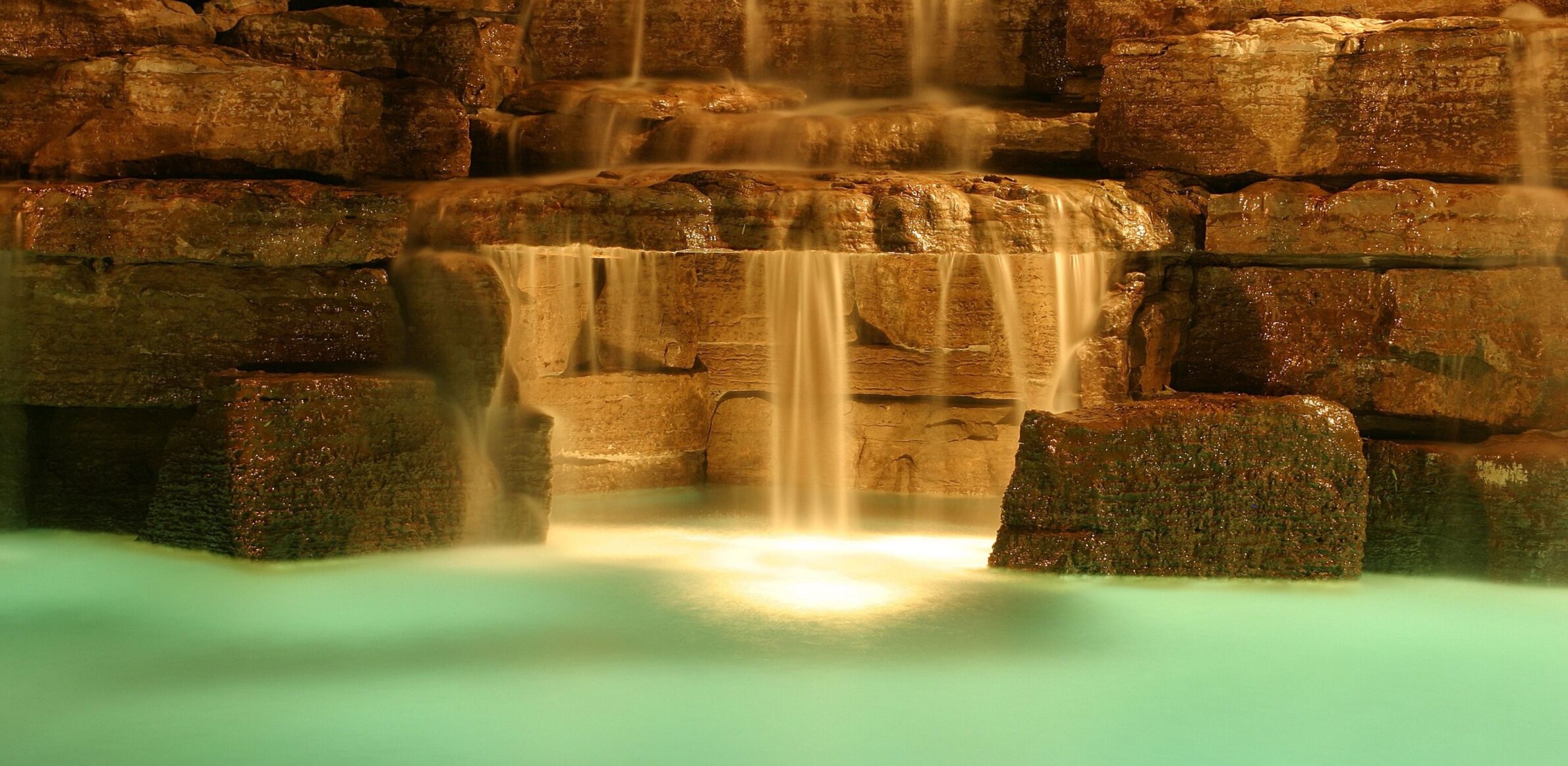 A serene, artificial waterfall cascades over mossy, illuminated rocks into a tranquil turquoise pool, likely in a garden or indoor setting.