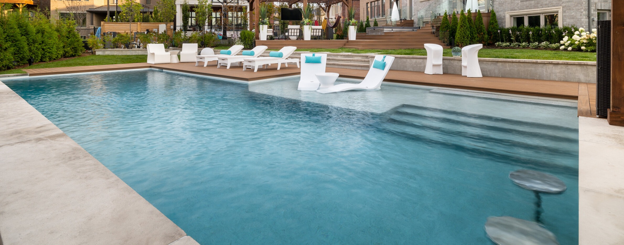 An outdoor swimming pool with crystal clear blue water, surrounded by white lounge chairs, a wooden deck, and a well-manicured lawn in the background.