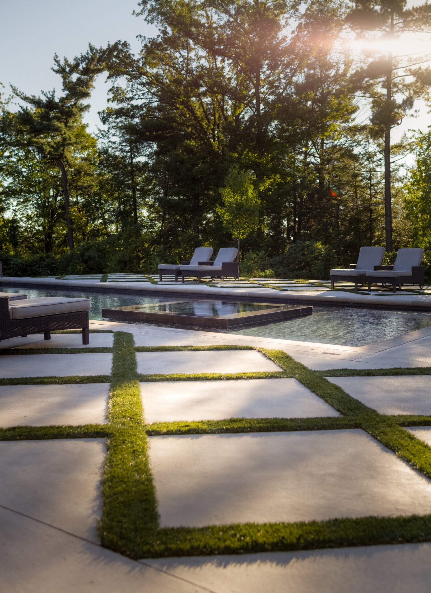 This image shows an outdoor swimming pool surrounded by trees at sunset, with lounge chairs, a woven sofa, and neat grass-bordered stone pathways.