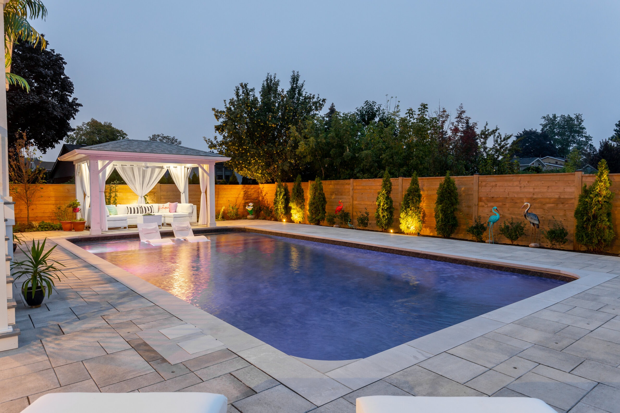 An elegant outdoor swimming pool at dusk, with a white gazebo, lounge chairs, ambient lighting, decorative bird sculptures, and a landscaped garden.