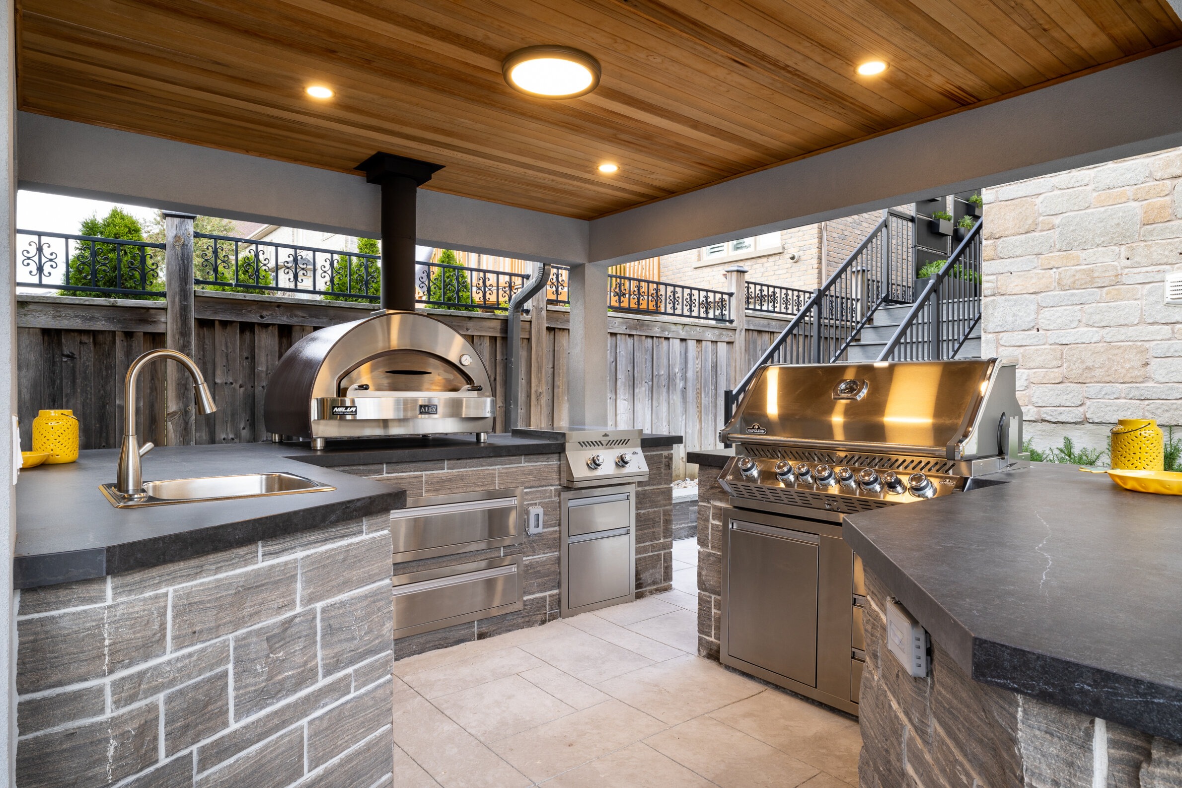 Outdoor kitchen with stainless steel appliances, pizza oven, grill, sink, and storage, under a wooden ceiling, next to a staircase and stone wall.
