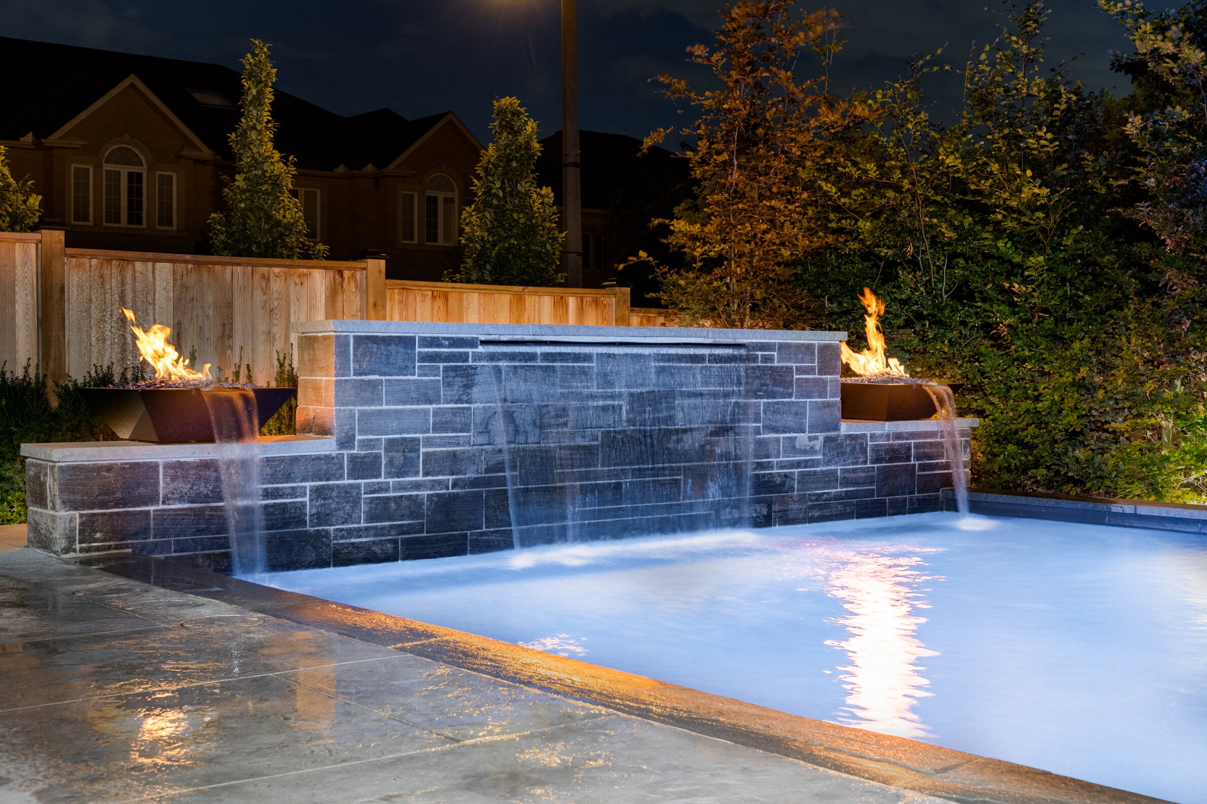 An outdoor pool with a dark stone waterfall feature, and two fire pits glowing at night, surrounded by wooden fencing and greenery.