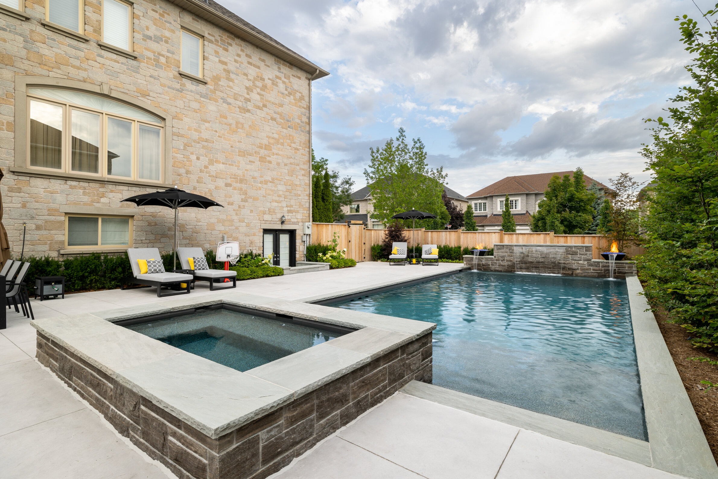 A luxurious backyard with a swimming pool, hot tub, sun loungers, outdoor furniture, a fireplace, and a stone house under a cloudy sky.