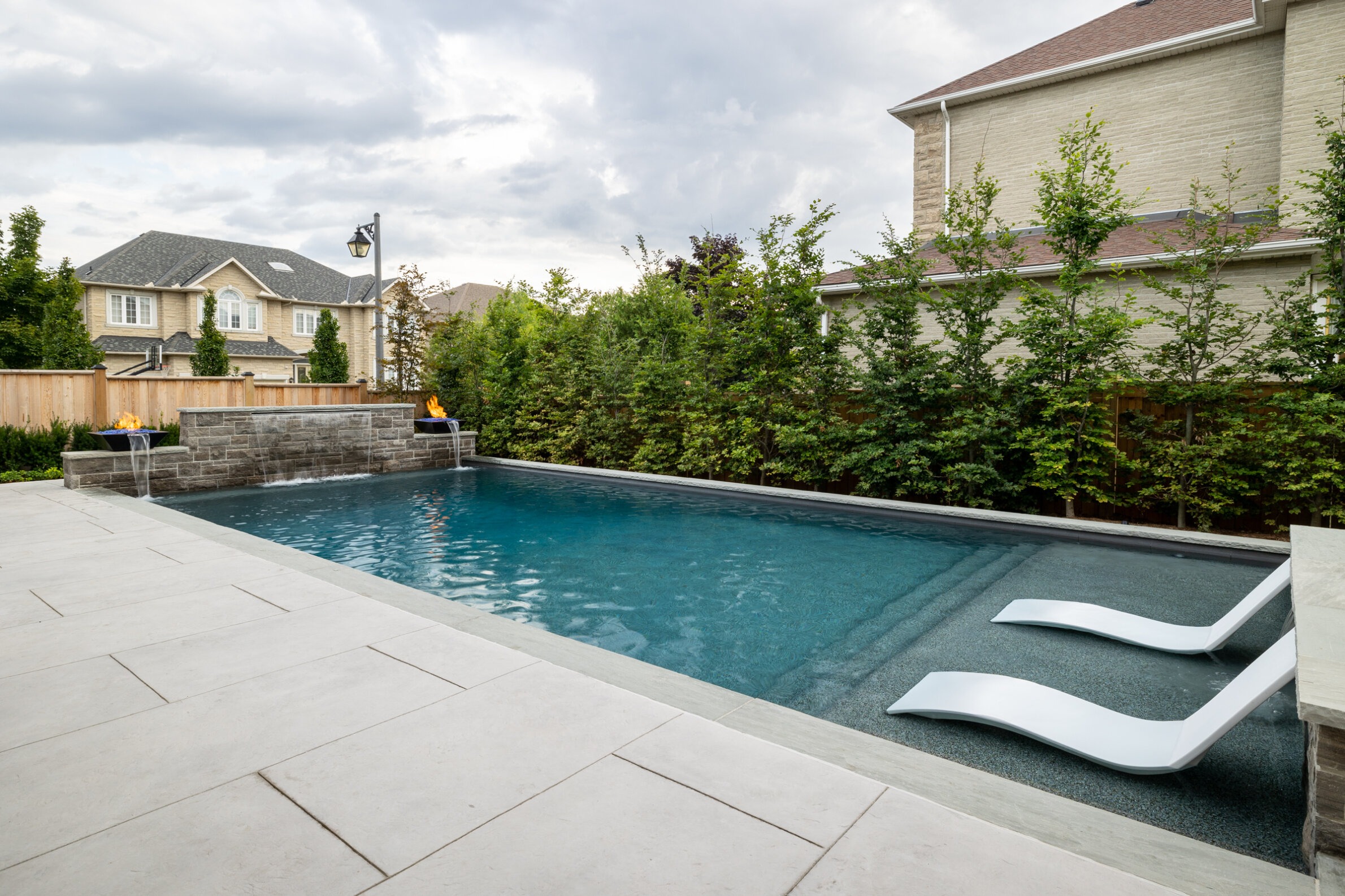 Residential backyard with a rectangular swimming pool, two modern white loungers, stone wall with fire features, and a lush green fence line.