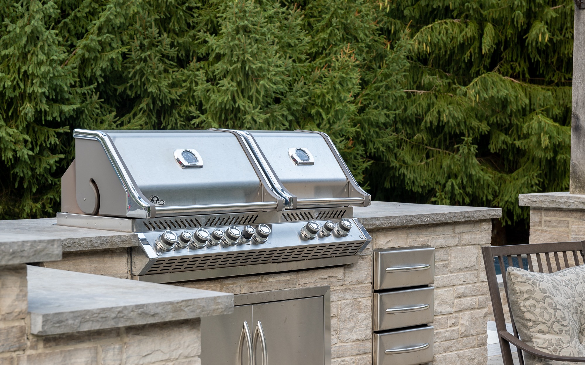An outdoor kitchen with two stainless steel grills built into a stone counter, with evergreen trees in the background and patio furniture nearby.