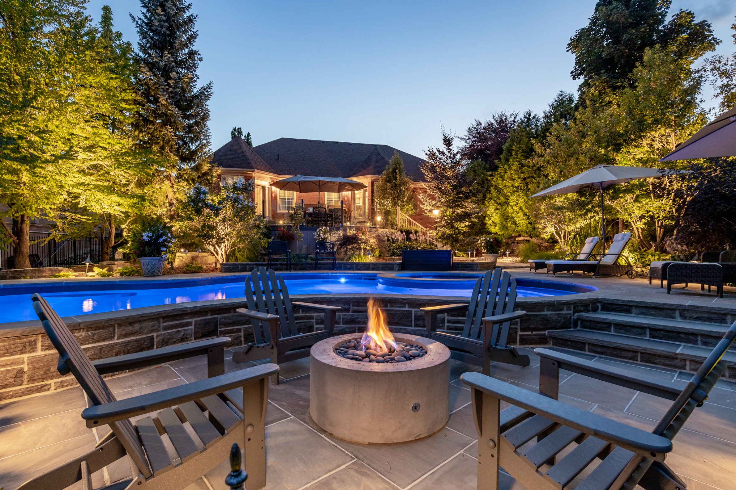 A luxurious backyard at dusk with a lit swimming pool, fire pit, outdoor chairs, lush trees, and a house in the background.