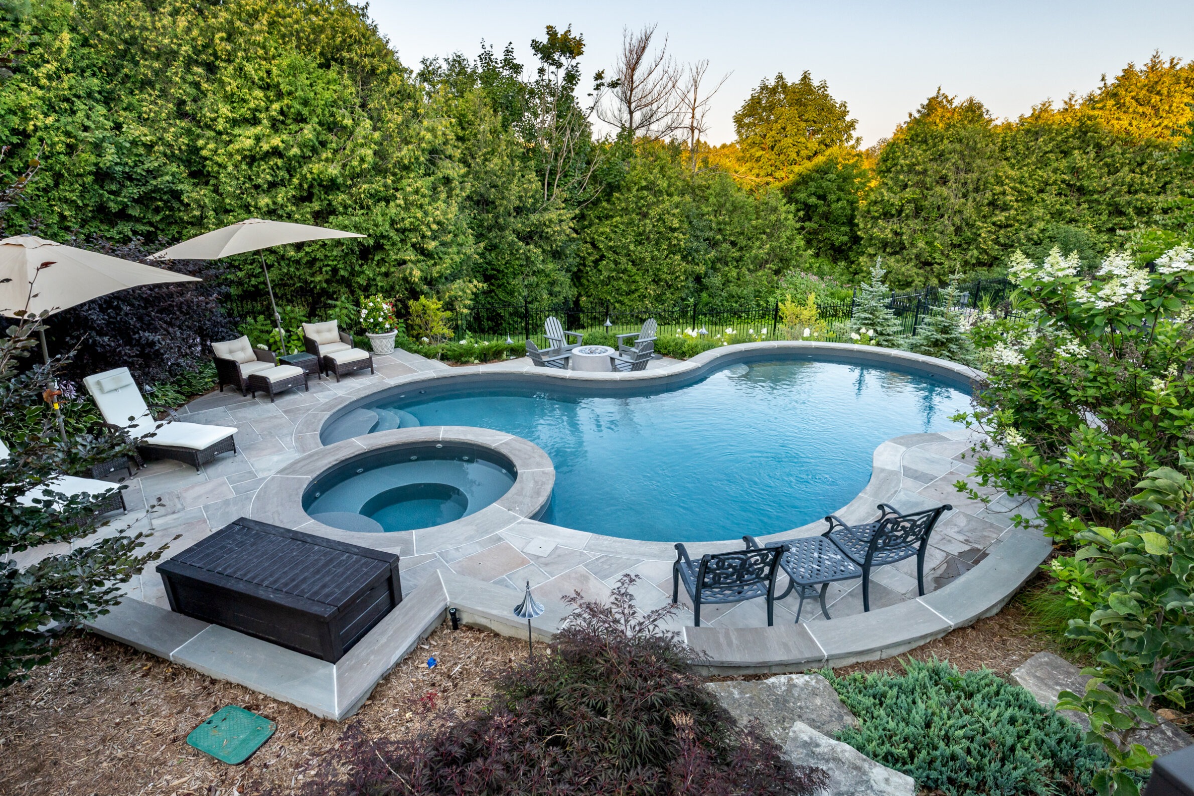 This image shows an outdoor residential swimming pool with a hot tub, surrounded by patio furniture, greenery, and a clear blue sky above.