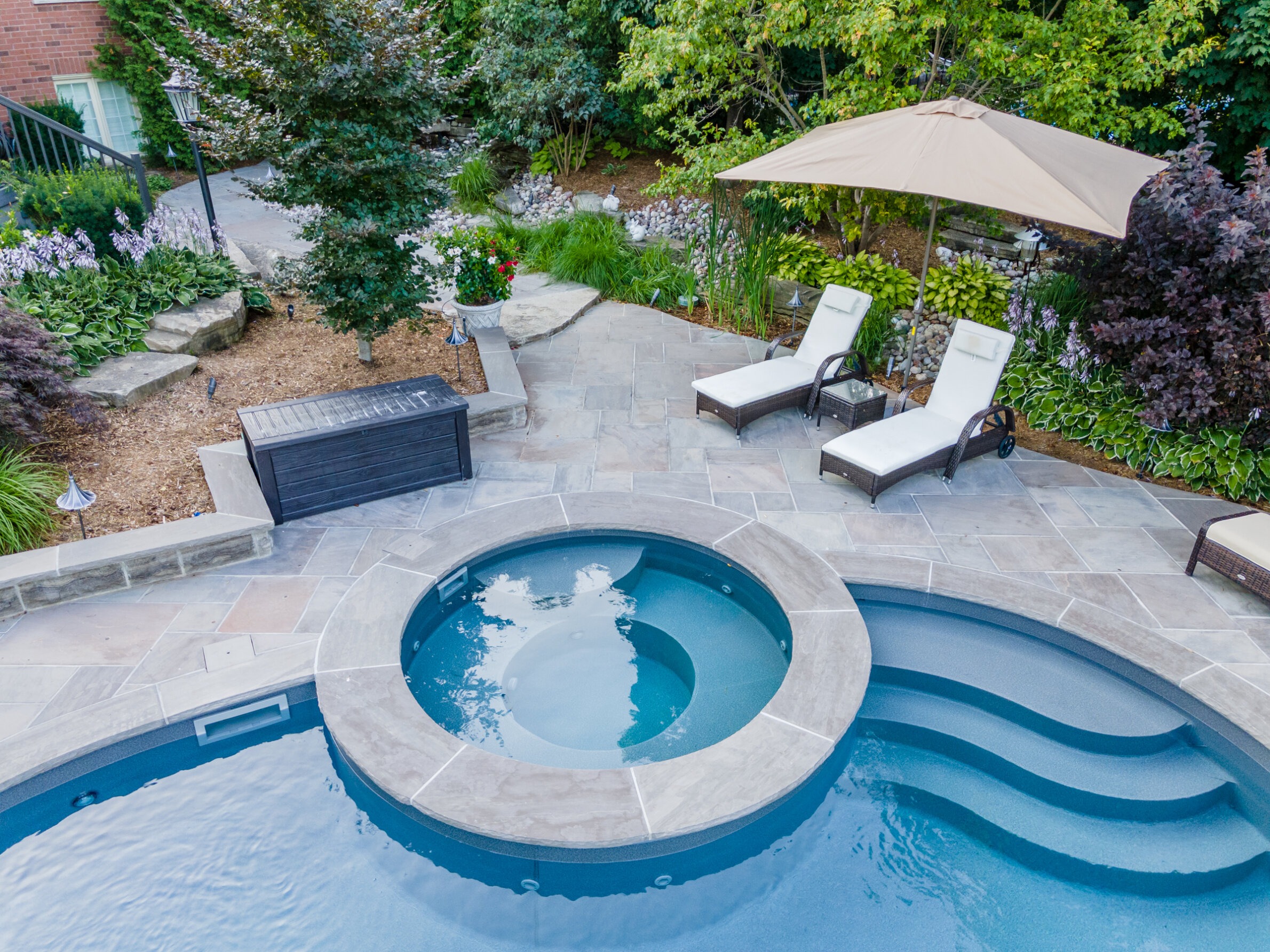 An aerial view of a backyard with a curved pool, hot tub, two lounge chairs under an umbrella, surrounding landscaped garden, and a storage box.