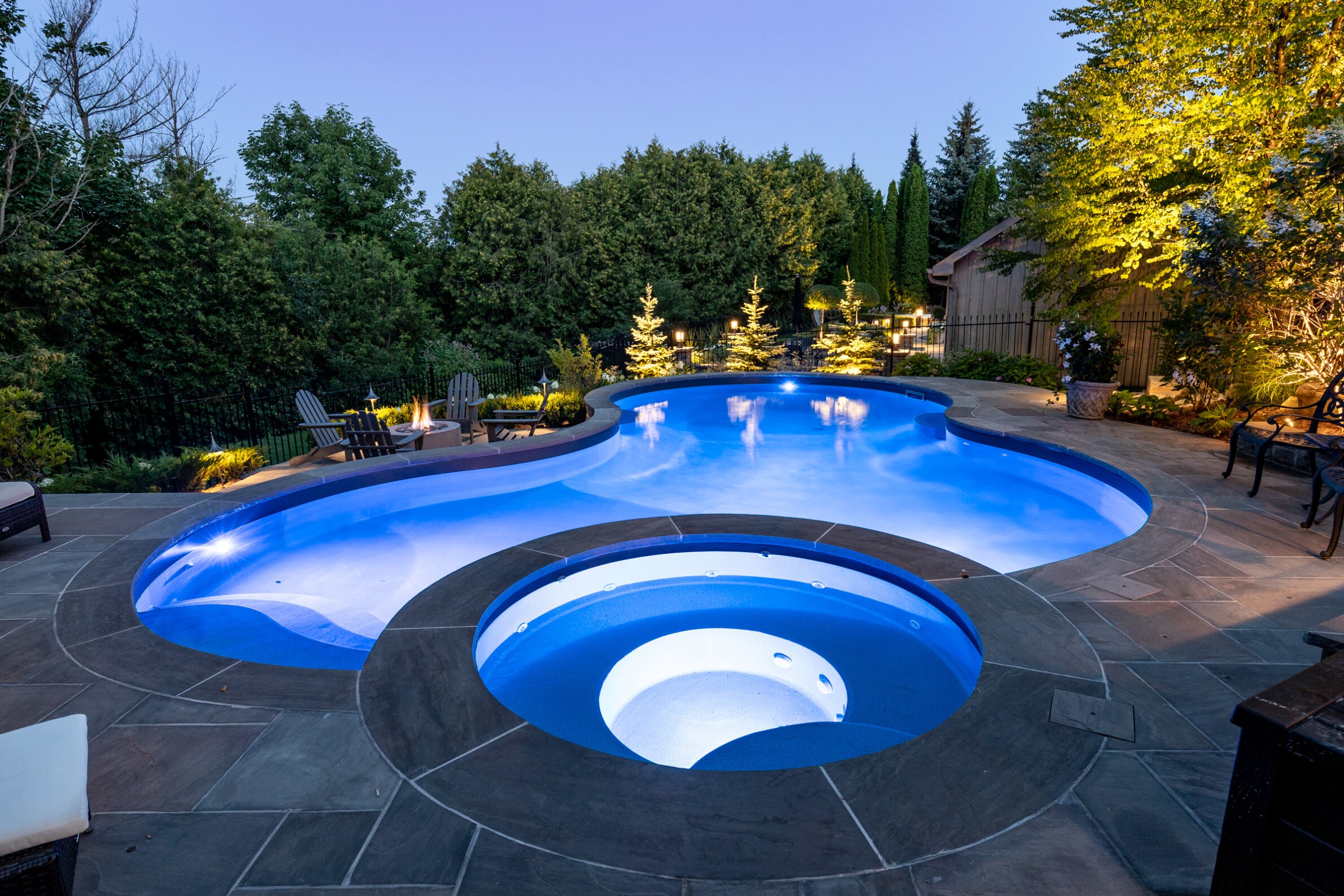 A luxurious backyard pool with blue lighting, adjacent hot tub, surrounded by lush trees and equipped with patio furniture and a lit fire pit.