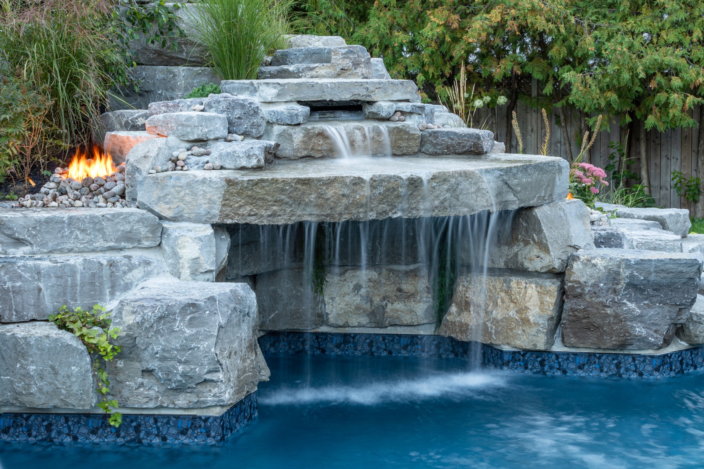An artificial cascading waterfall built with layered stone flows into a pool with clear blue water. A fire pit emits flames near greenery and a fence.