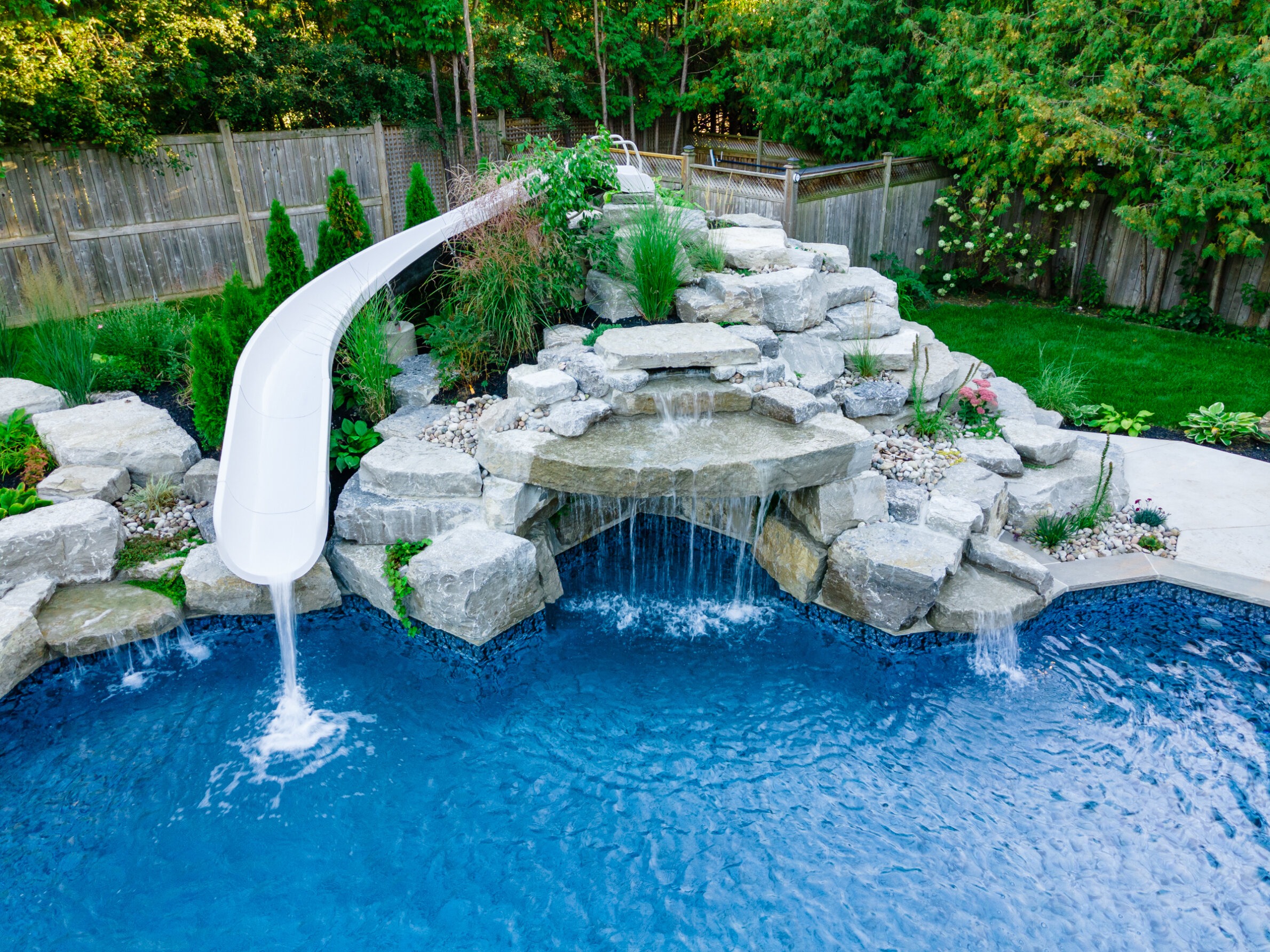 An outdoor pool with a white slide and a rock waterfall feature surrounded by a landscaped garden, lawn, and a wooden fence.