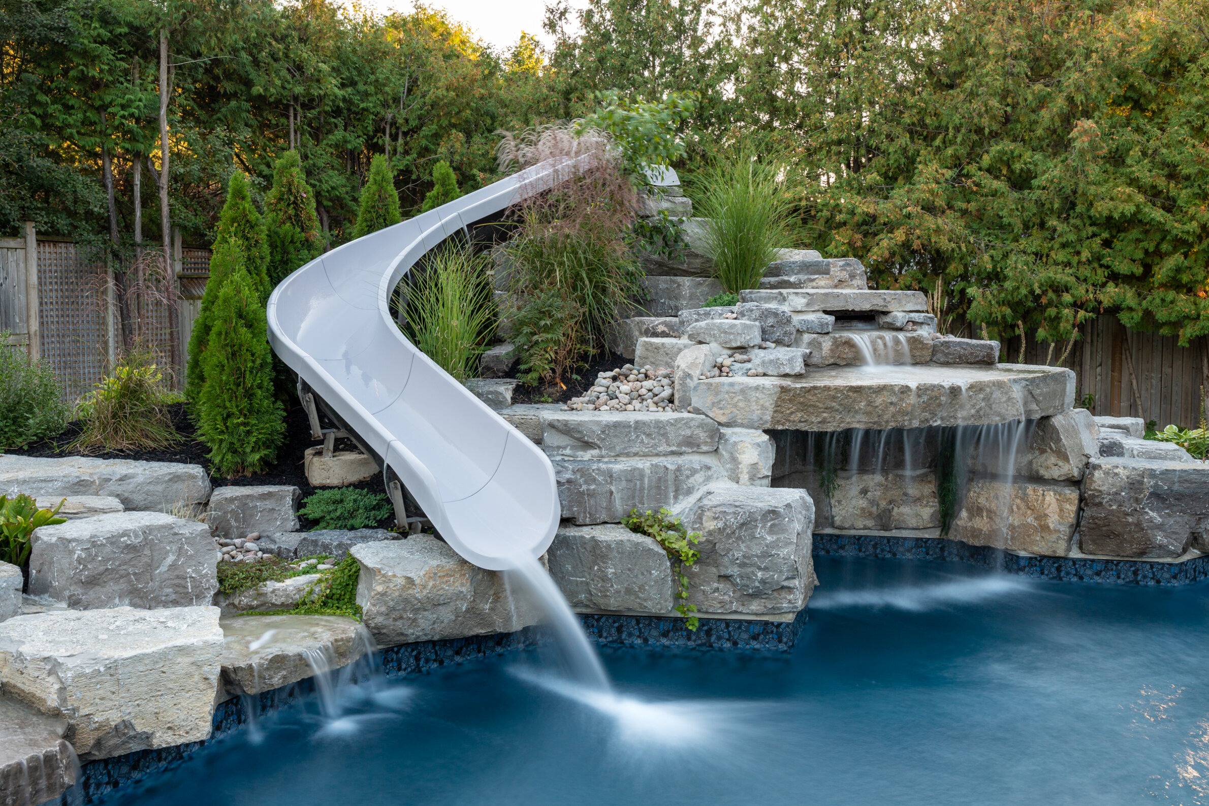This image features a residential backyard with a pool, a curved water slide, a rock waterfall, landscaped plants, and a wooden fence.