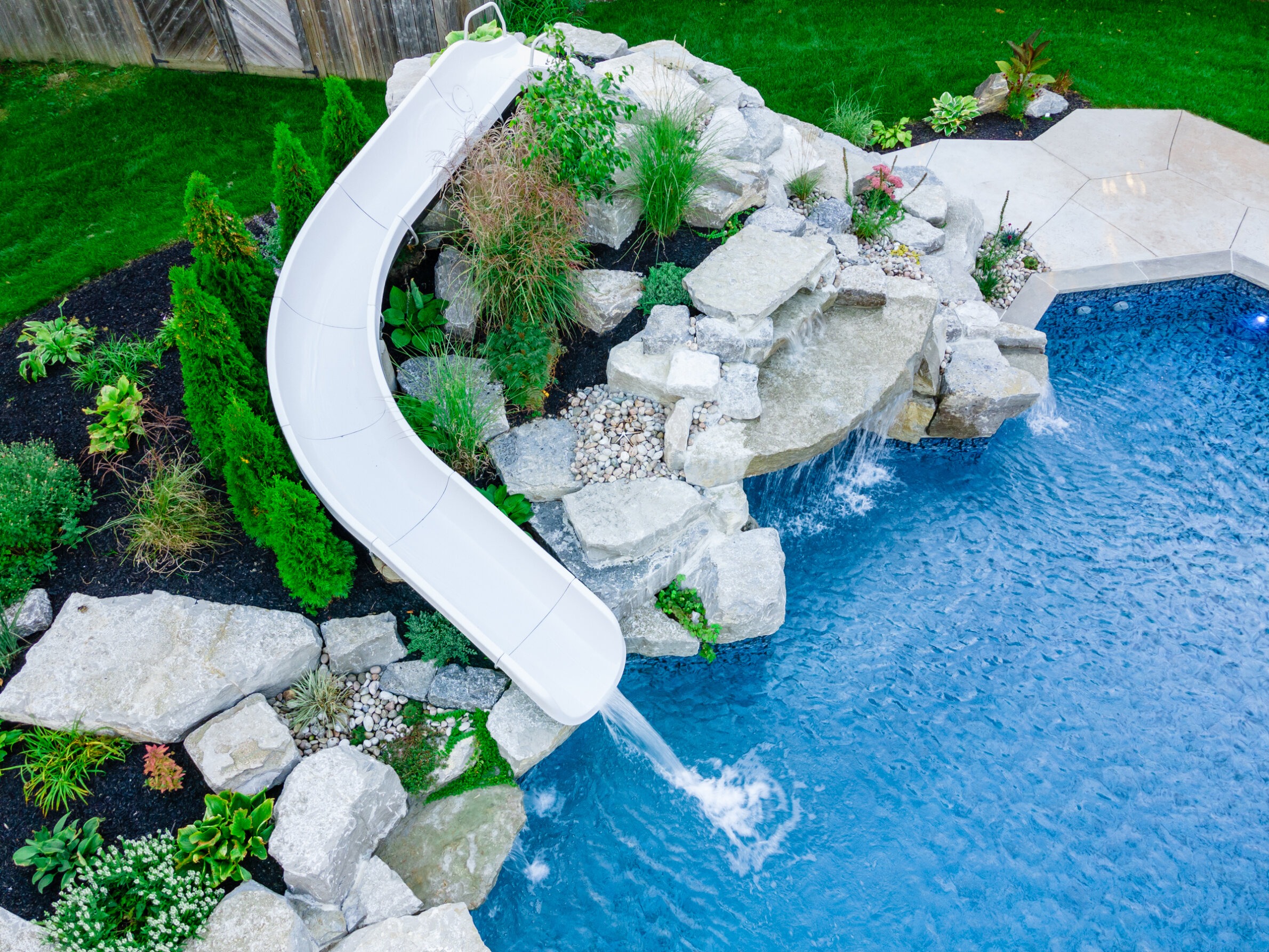 An aerial view of a backyard with a white slide descending into a blue swimming pool, surrounded by rocks and landscaping with green plants.