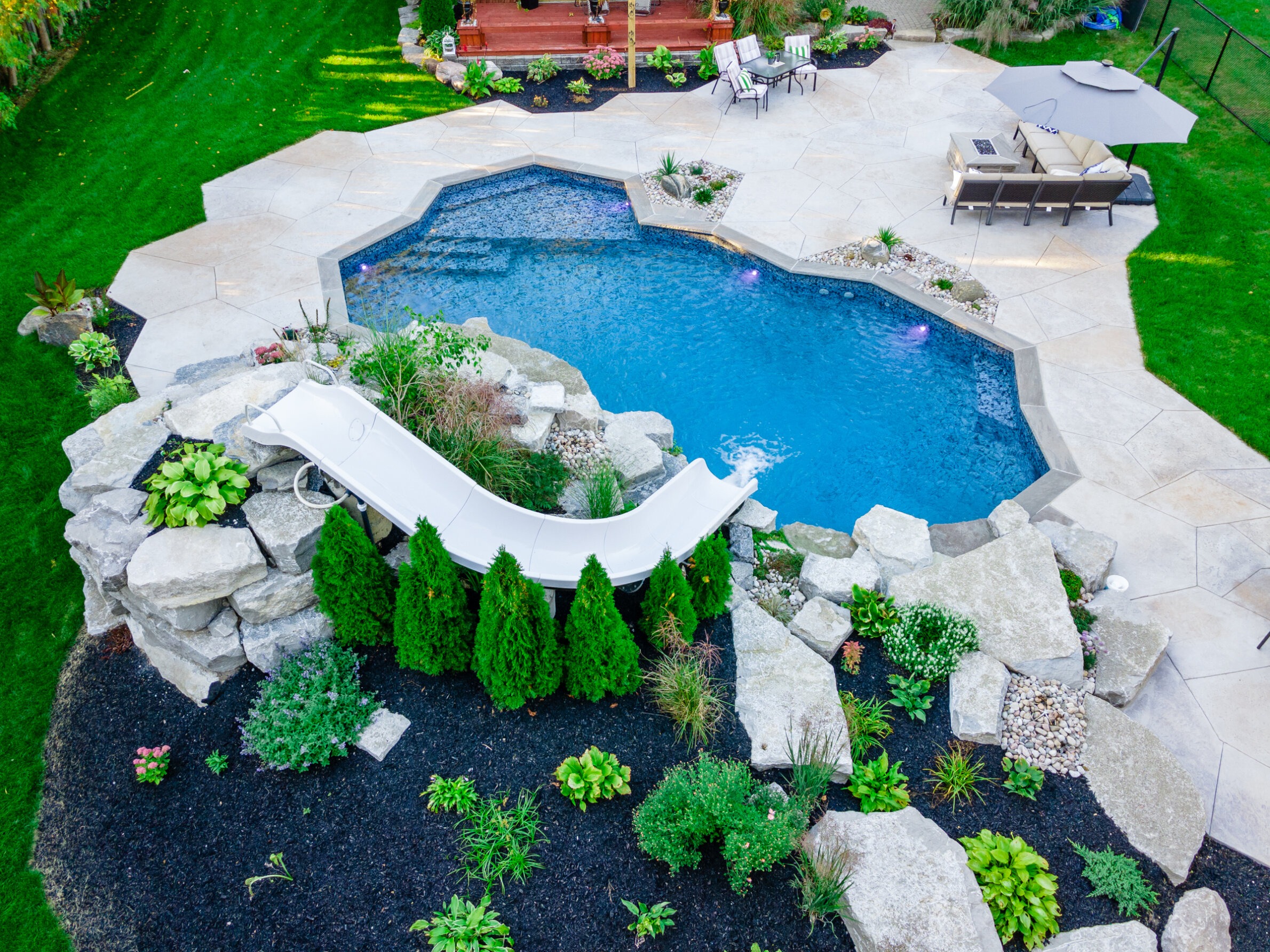 Aerial view of a landscaped backyard featuring an irregularly shaped swimming pool with slide, surrounded by stone patio, greenery, and outdoor furniture.