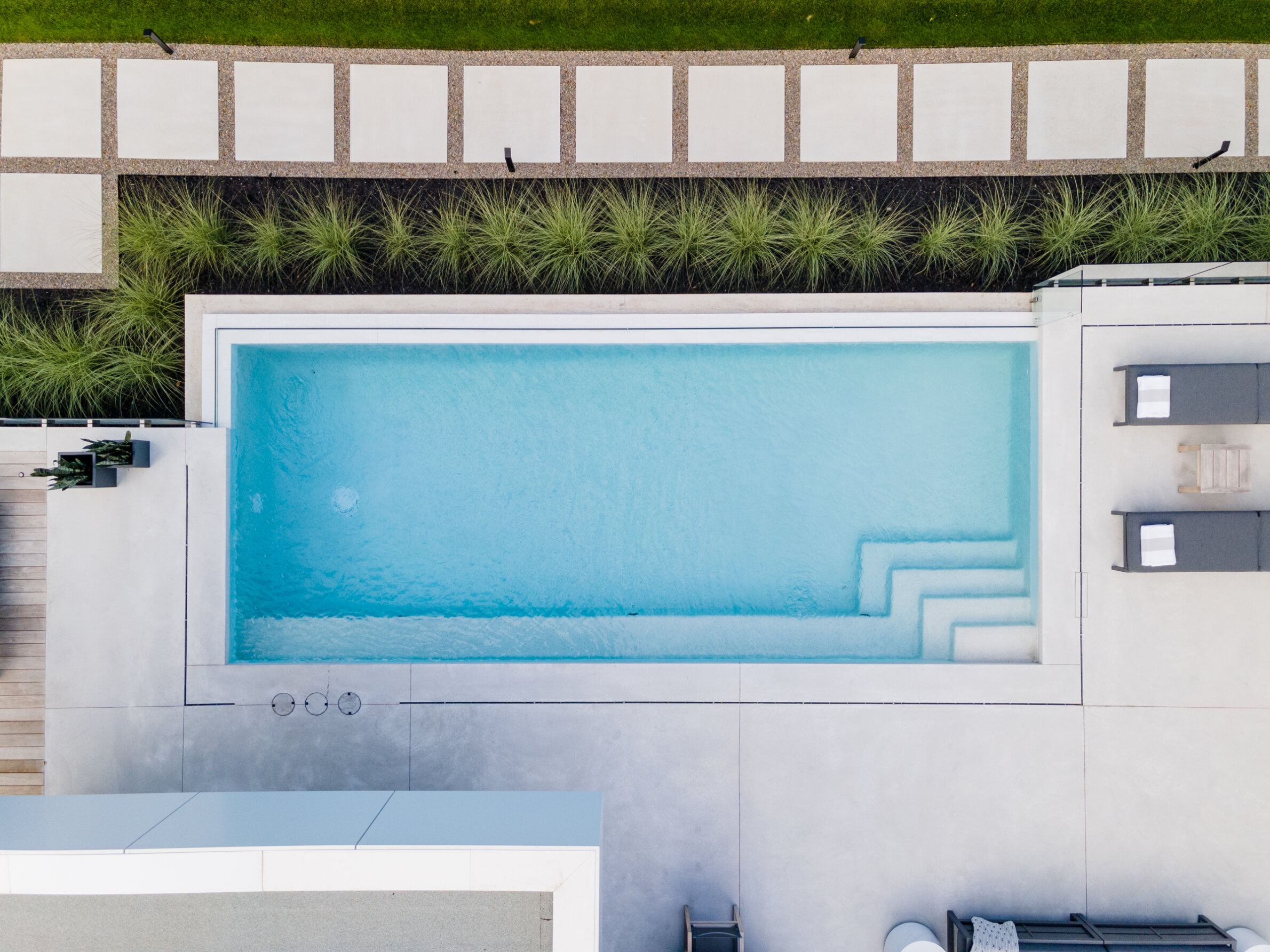Aerial view of a modern backyard with a rectangular swimming pool, concrete patio, neatly arranged pavers, and ornamental grass landscaping.