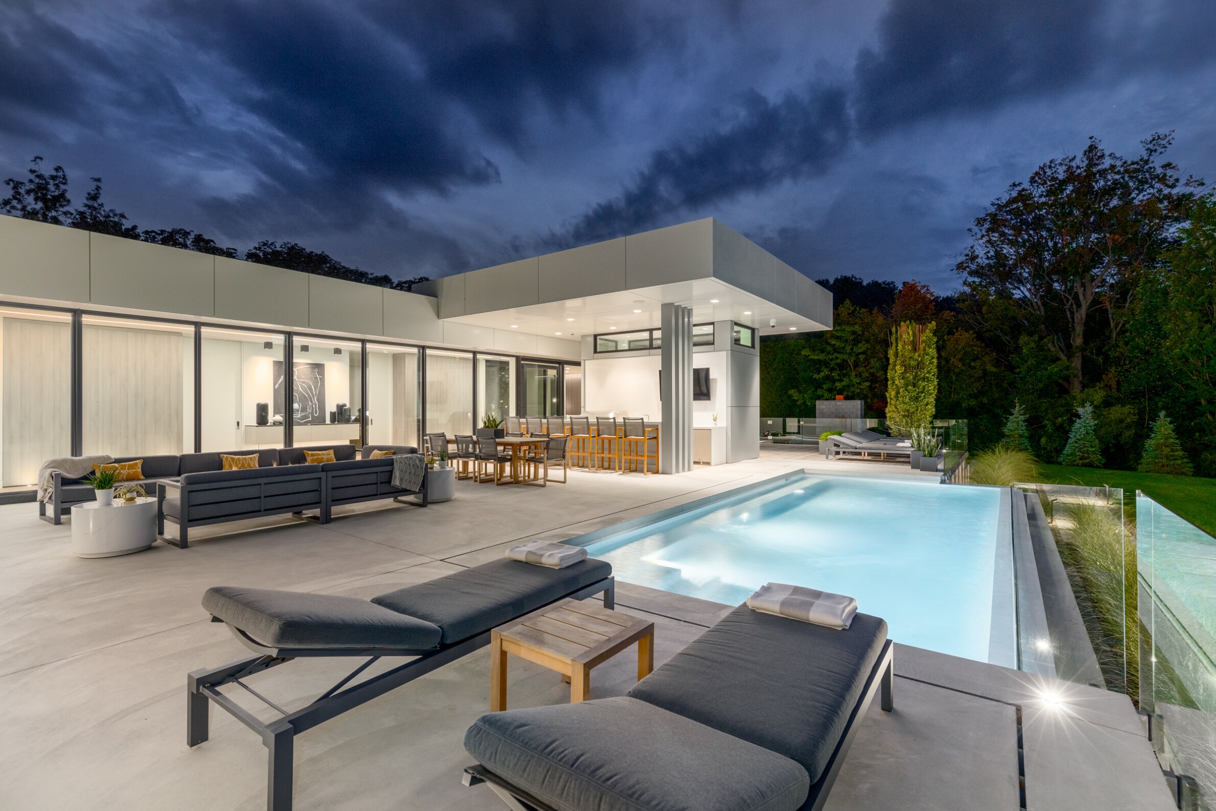 A modern house with large windows and an outdoor pool at dusk. Patio with loungers and an outdoor dining area, surrounded by trees under a dramatic sky.