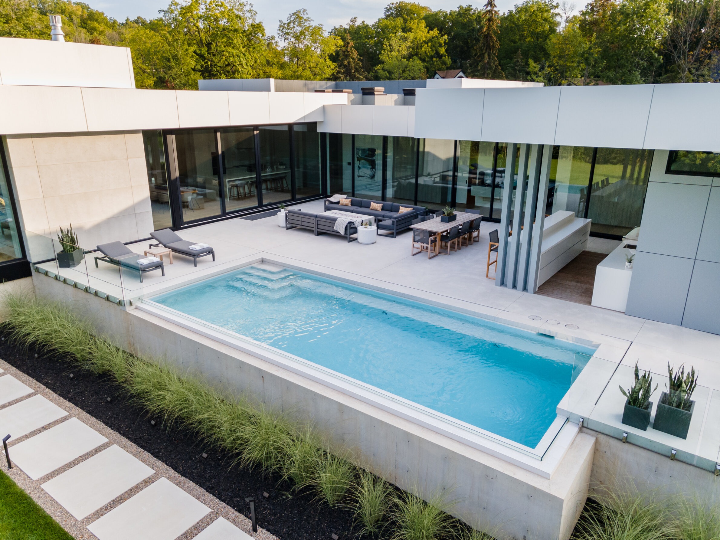 Modern house with large windows, outdoor furniture on patio, rectangular swimming pool, and landscaped garden. Luxurious setting with contemporary design.