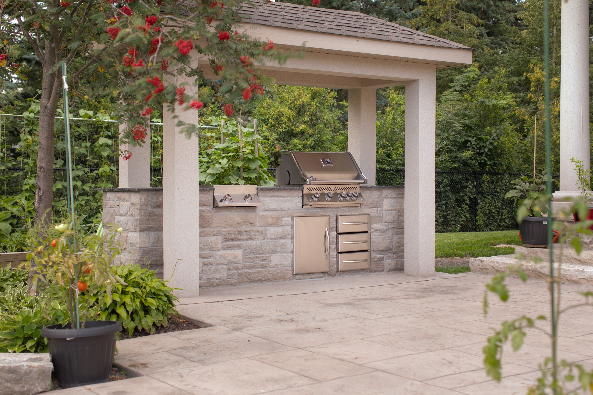 An outdoor kitchen pavilion with a built-in stainless steel grill, storage drawers, under a shelter, surrounded by green plants, and red flowers.