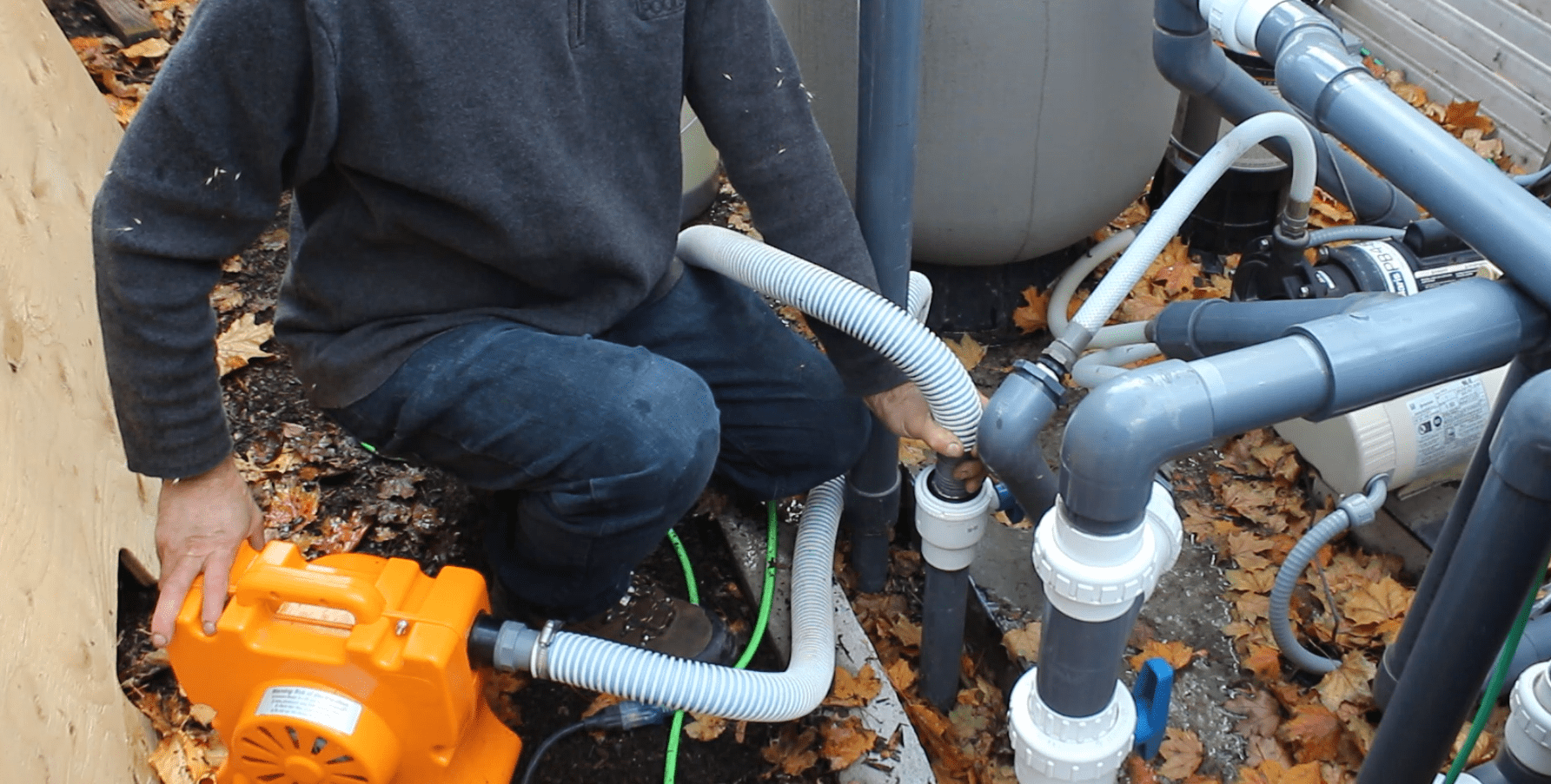 A person is kneeling beside a pool pump and filtration system, surrounded by fallen leaves, adjusting equipment outdoors.