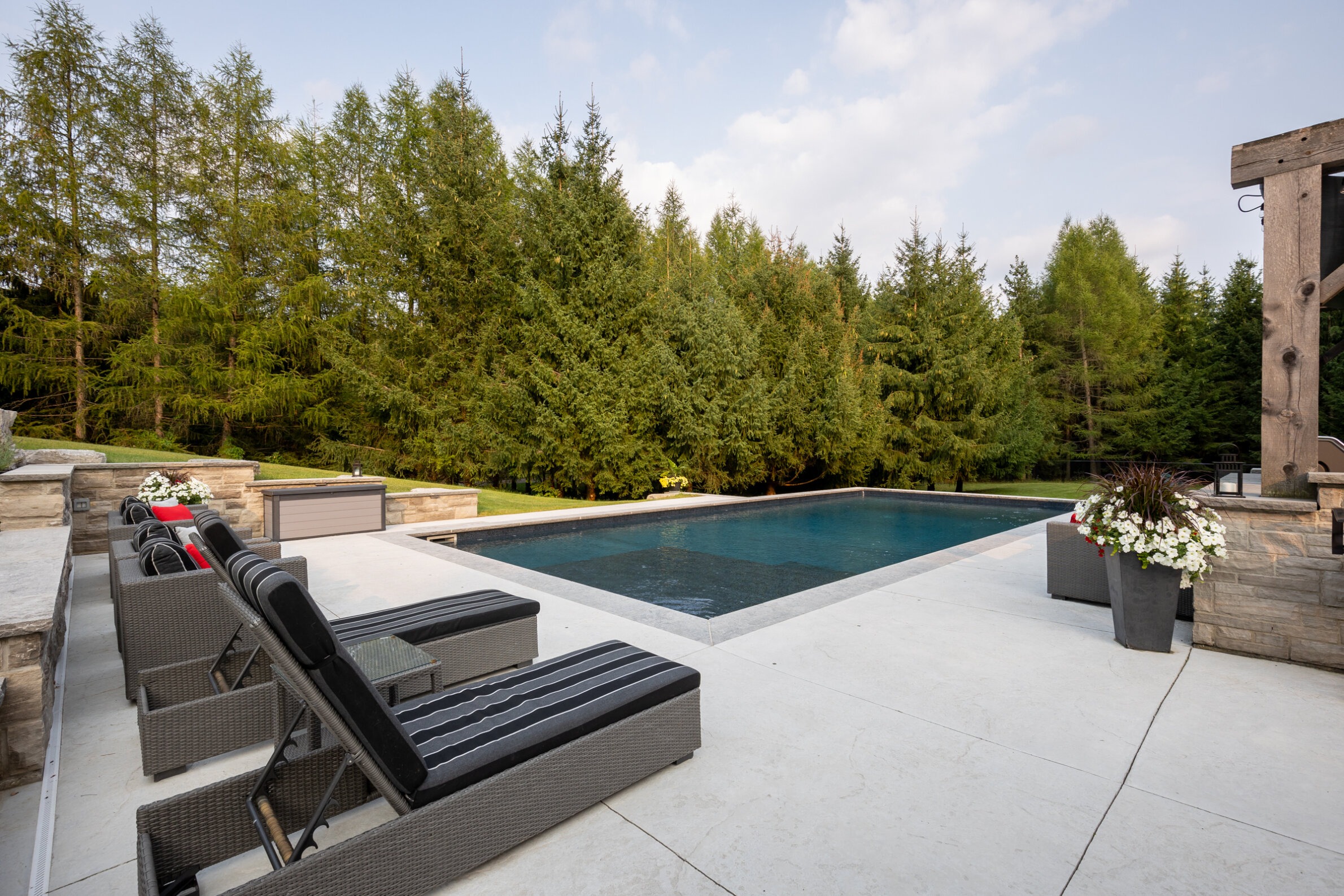 Luxurious outdoor pool with dark water, surrounded by trees, featuring lounge chairs, a fire pit, and a clear sky at dusk or dawn.