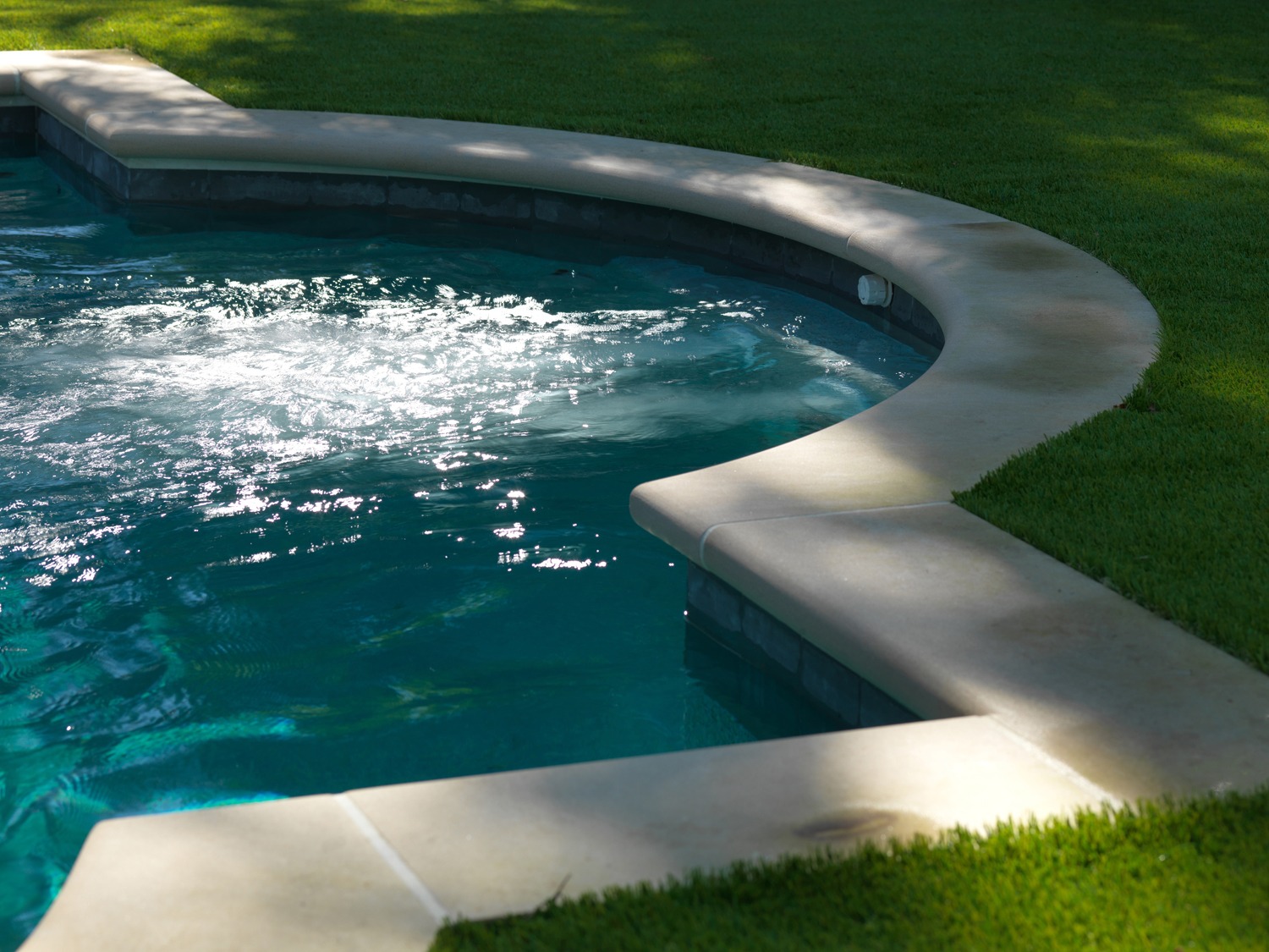 An outdoor swimming pool with sparkling water is surrounded by a curved, sunlit patio and neatly trimmed grass, suggestive of a tranquil backyard setting.
