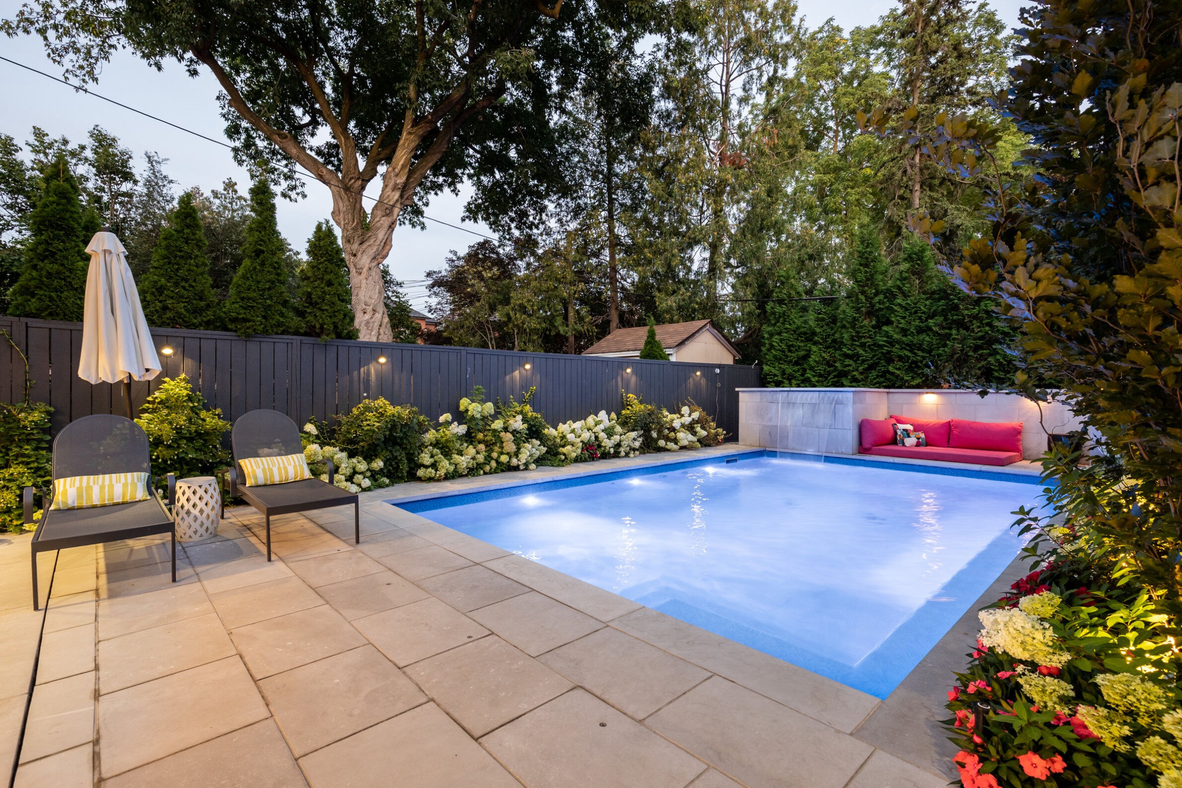 The image depicts a serene, illuminated backyard with an in-ground pool, patio, loungers, flowers, and a seating area under a large tree at dusk.