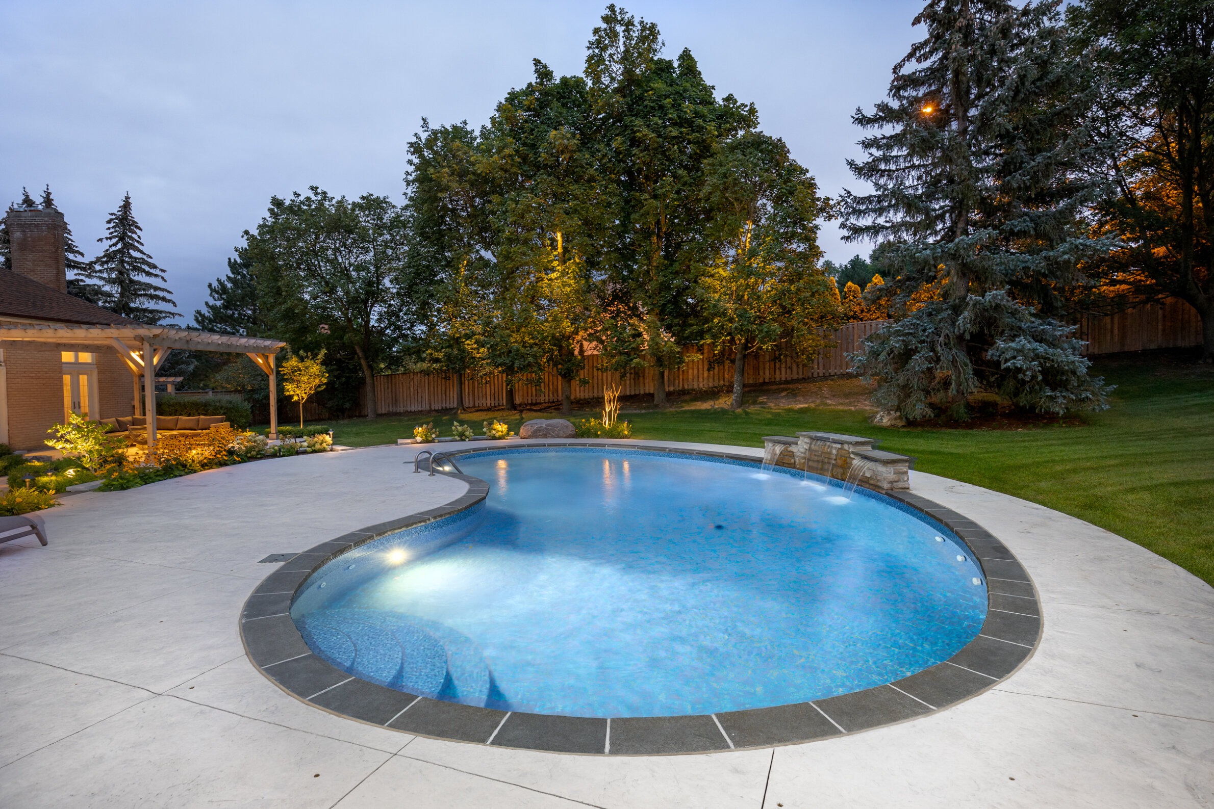 An elegantly lit backyard at twilight, featuring a curved in-ground pool with a waterfall, surrounded by manicured grass and a perimeter fence.