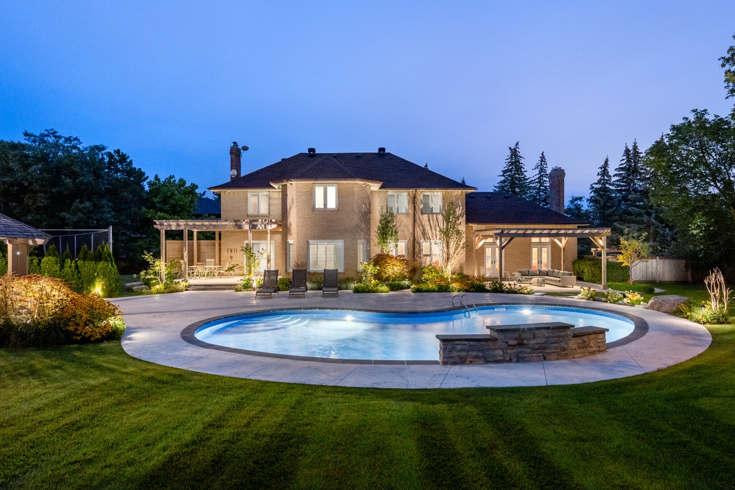 A luxurious two-story brick house at twilight with a lit swimming pool, landscaped garden, outdoor seating, and ambient lighting, set against a blue evening sky.
