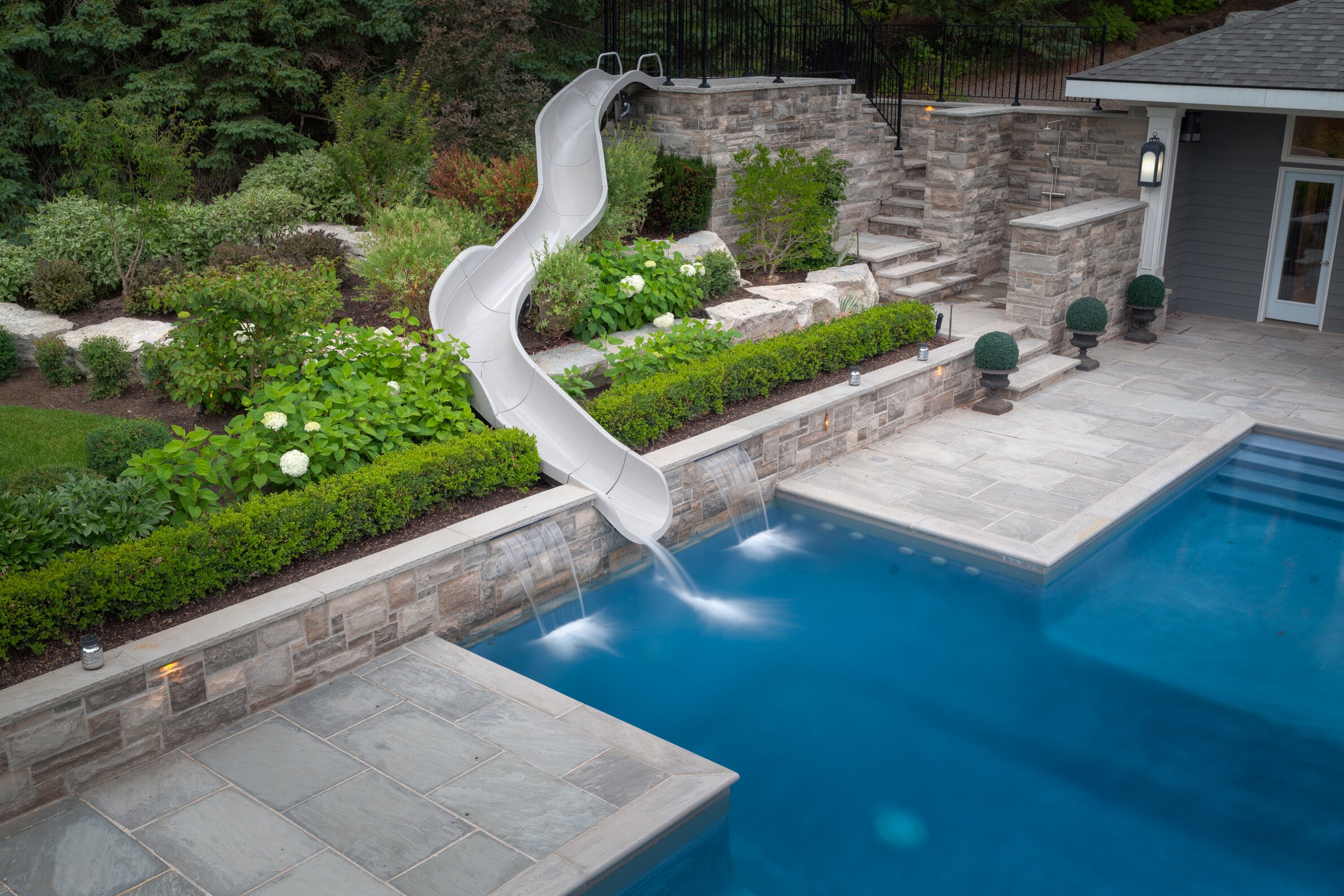 An outdoor residential swimming pool with a curved slide, landscaped garden, stone steps, and a house with exterior lighting visible in the background.