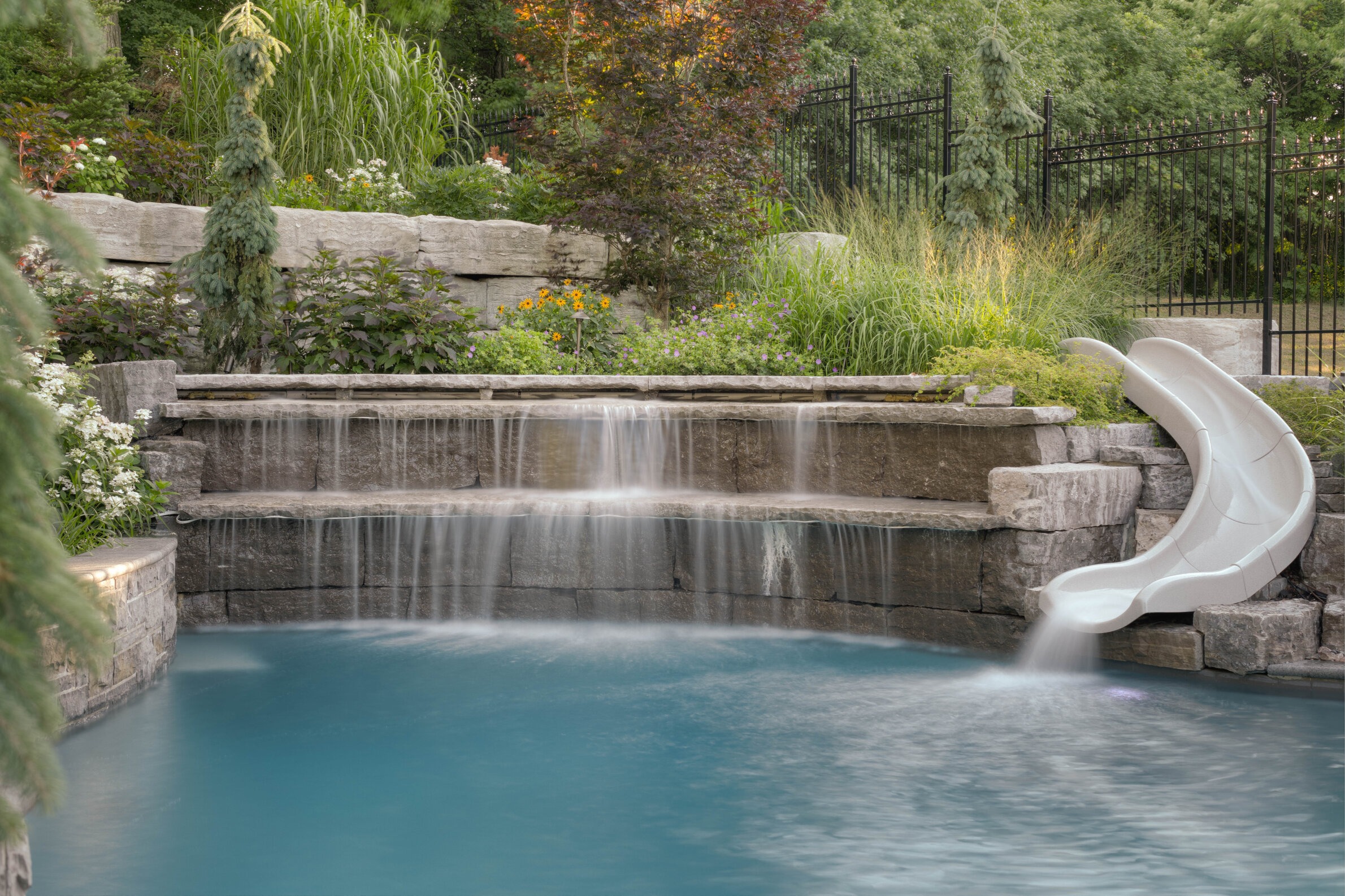 This image shows a tranquil backyard pool with a curved water slide and a cascading artificial rock waterfall set against lush greenery and flowers.