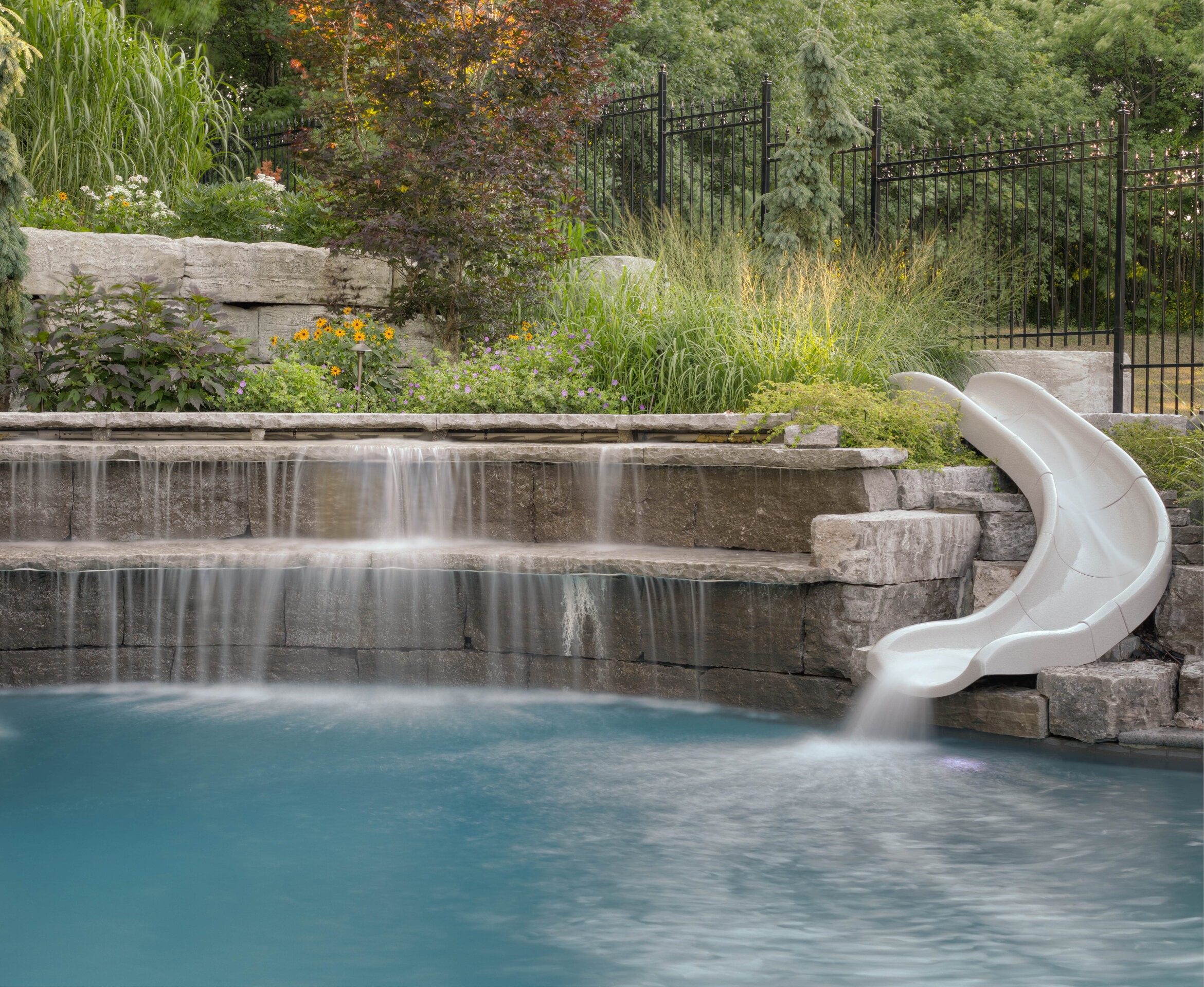 This image features a serene backyard with a swimming pool that includes a water slide and waterfall, surrounded by landscaped garden and a metal fence.