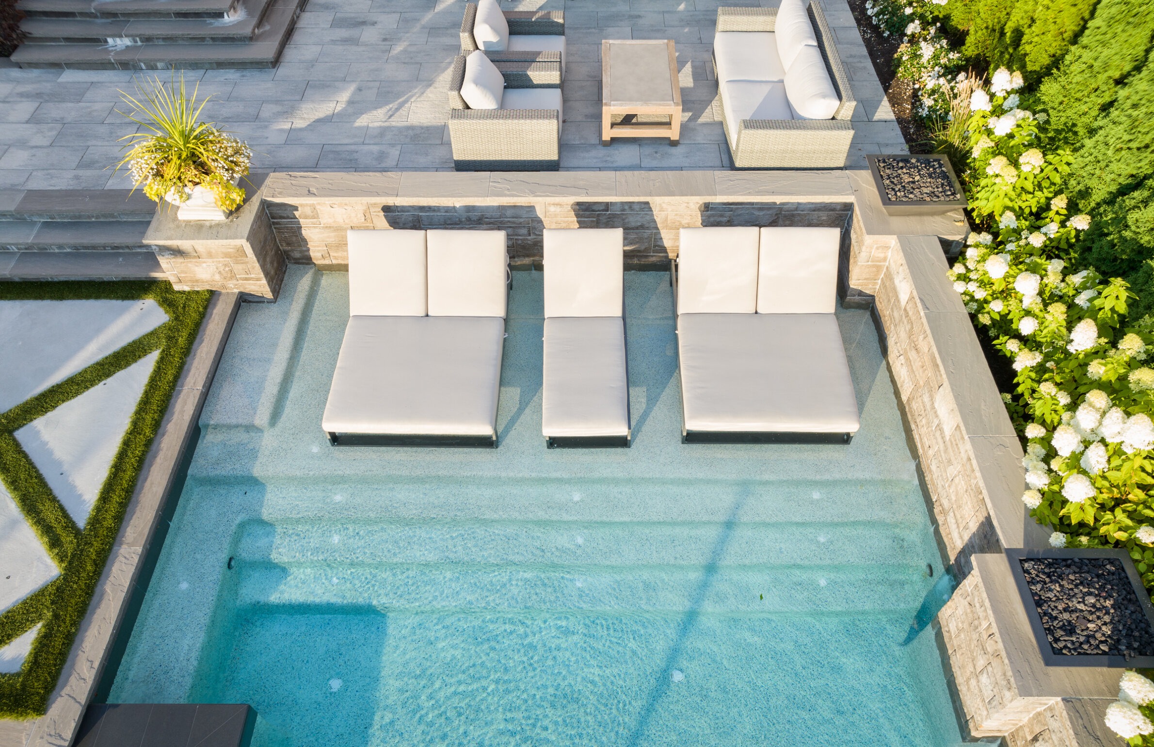 An aerial view shows a luxurious pool with clear blue water, surrounded by sun loungers, outdoor furniture, and lush greenery in a manicured garden.