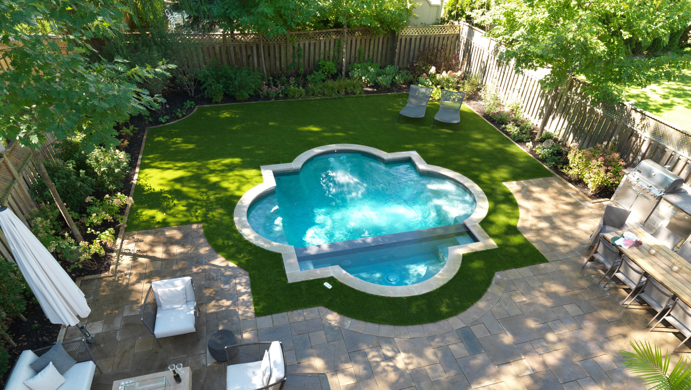 An aerial view of a cozy backyard featuring a kidney-shaped swimming pool, artificial turf, patio furniture, BBQ grill, and lush landscaping.
