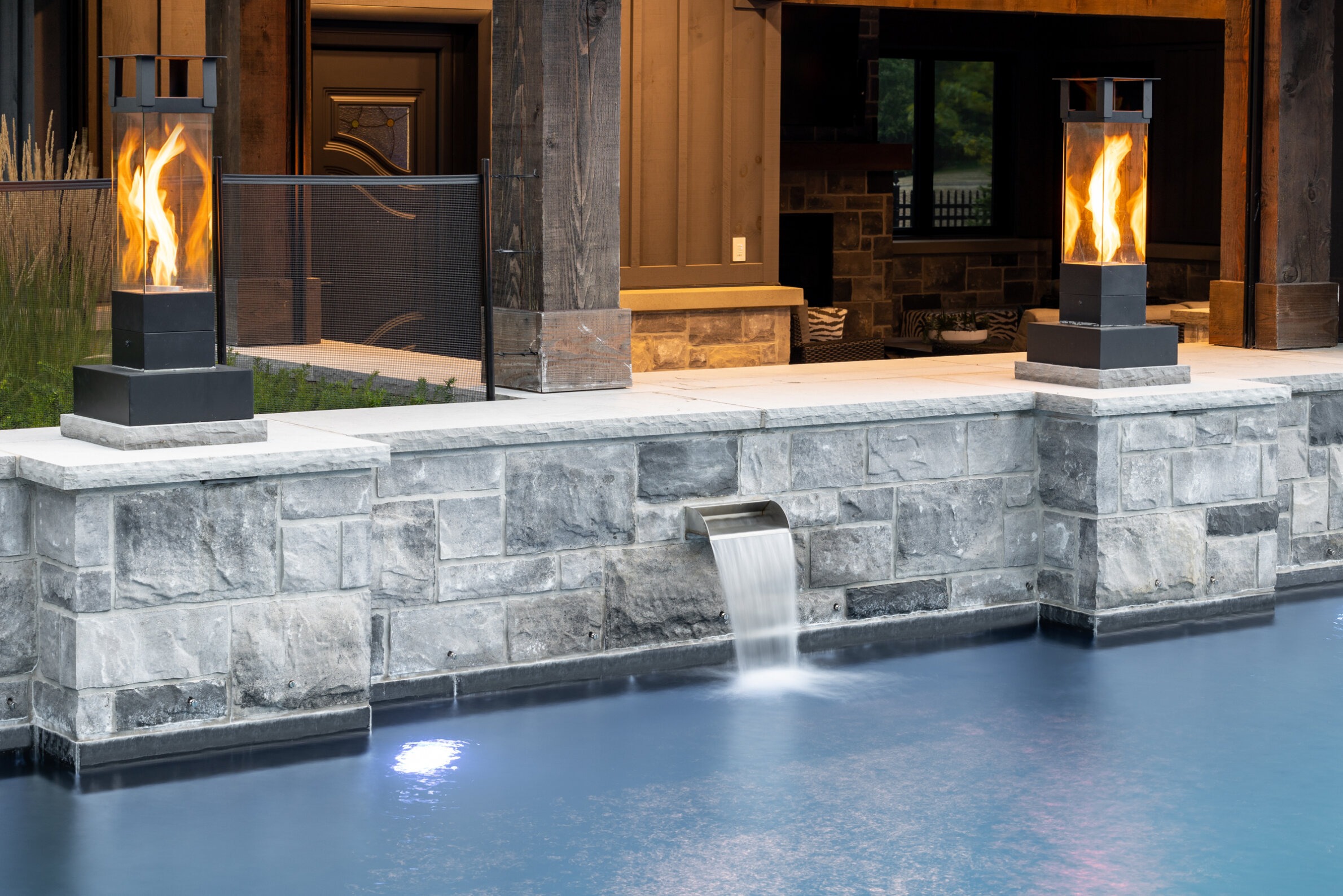 An outdoor patio features two flaming pillar torches beside a stone wall, overlooking a tranquil pool with a gently cascading water feature.