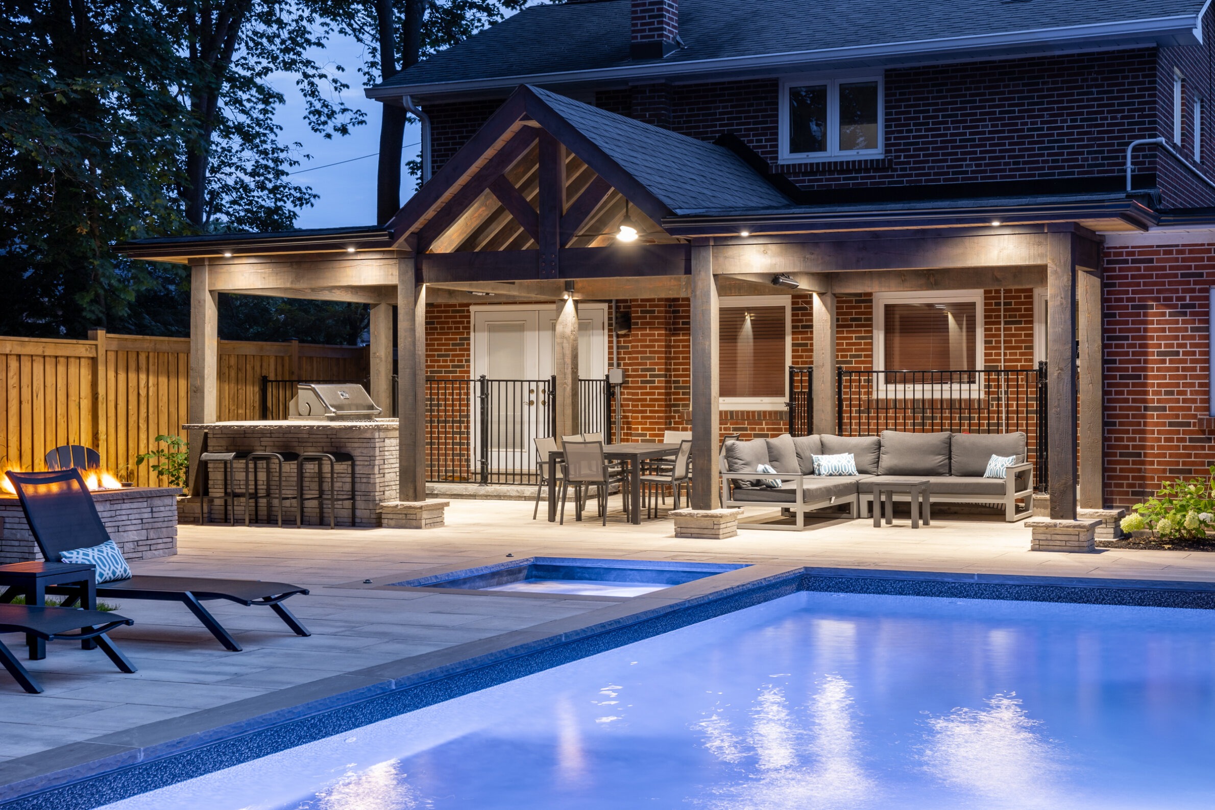 An outdoor residential space at dusk featuring a swimming pool, lit fire pit, lounge area, and a built-in grill beneath a covered patio.