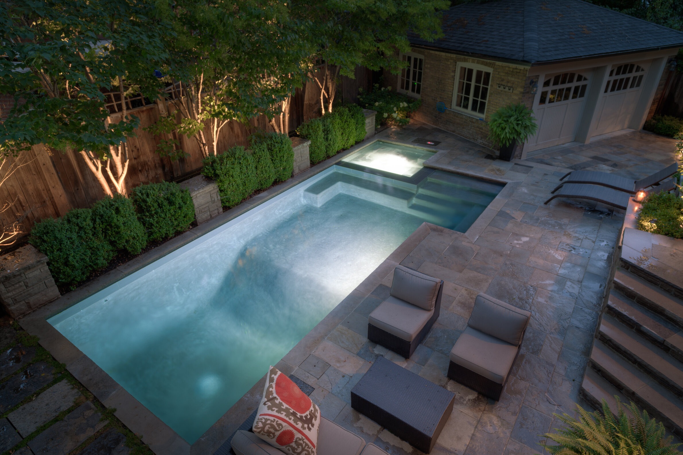 A serene backyard at dusk featuring a lit rectangular pool surrounded by lounging chairs, stone tiles, lush greenery, and a cozy pool house.