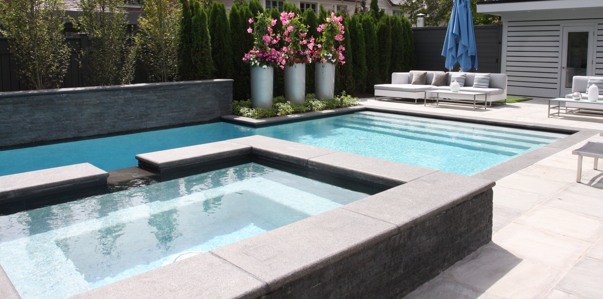 An elegant backyard with a blue swimming pool, surrounded by stone tile, large planters with pink flowers, lounge furniture, and a blue umbrella.
