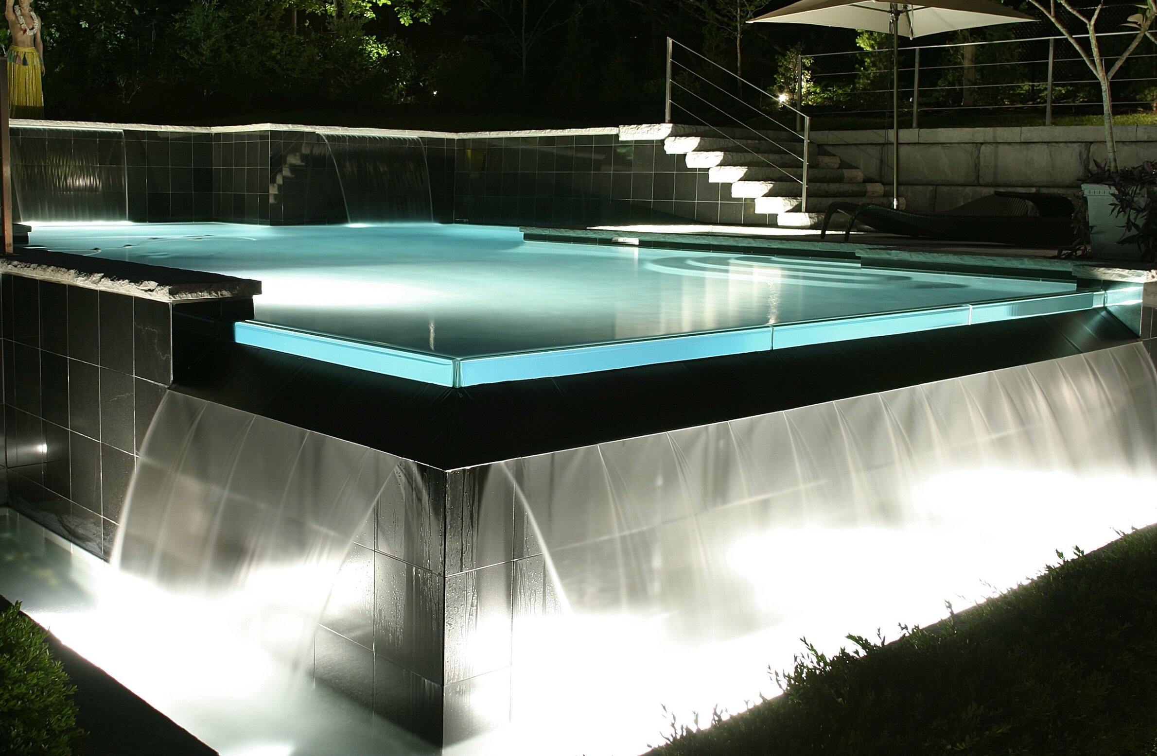 An illuminated outdoor swimming pool at night features cascading water, tiled edges, and steps with a stylish modern design and reflective surfaces.