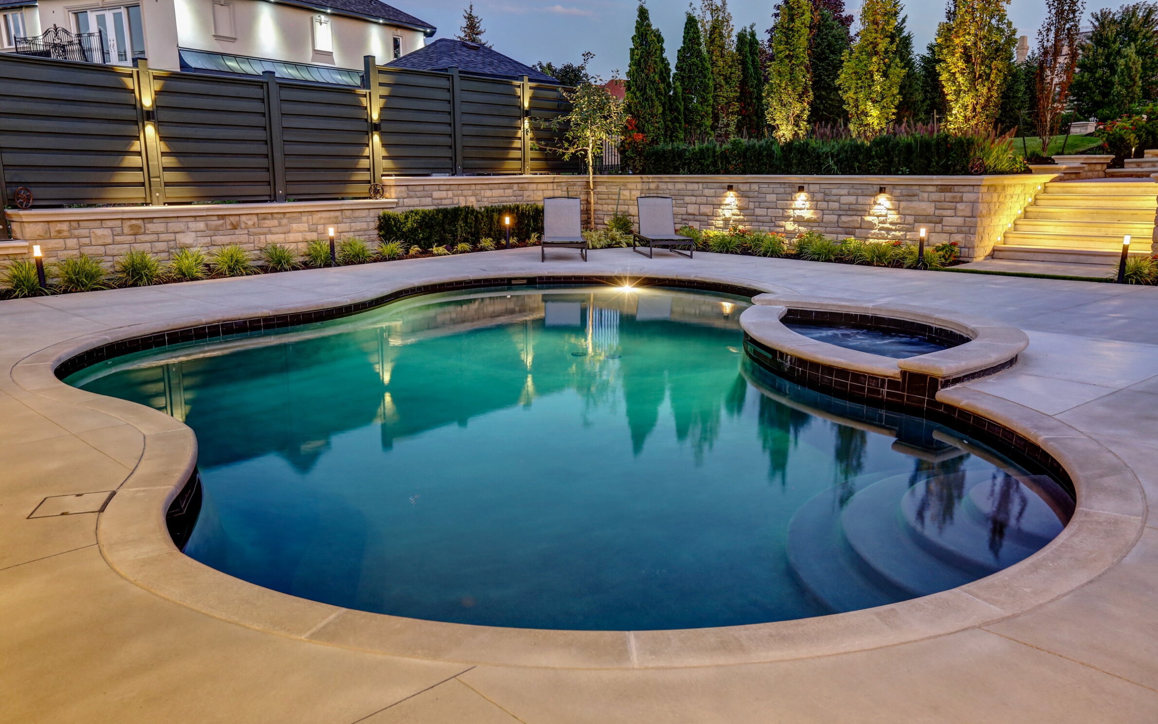 An outdoor swimming pool with an integrated hot tub, surrounded by stone landscaping, loungers, and garden lights, reflecting the evening sky.