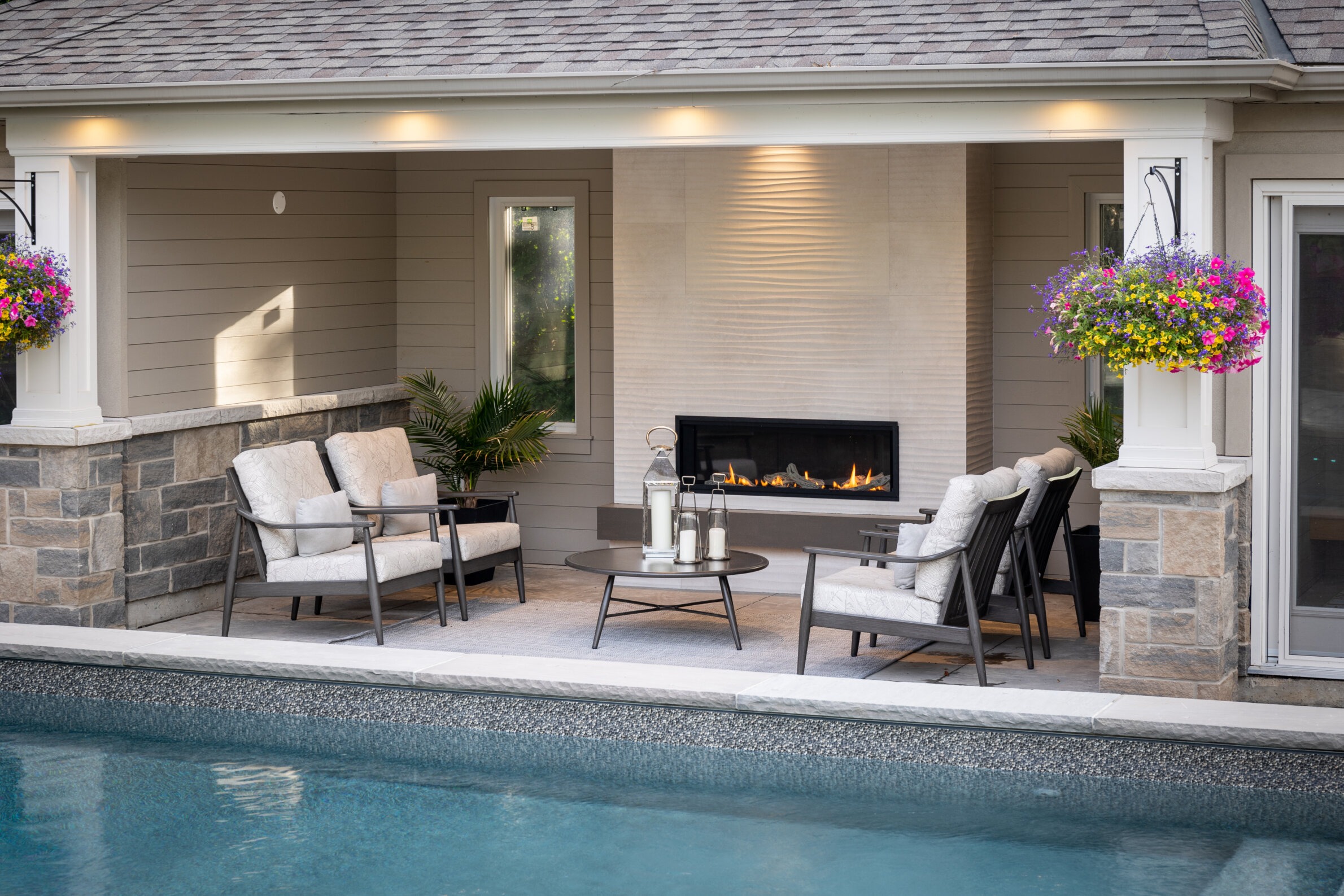 An outdoor patio with a fireplace, comfortable seating, and vibrant hanging flower baskets beside a swimming pool in a residential backyard setting.